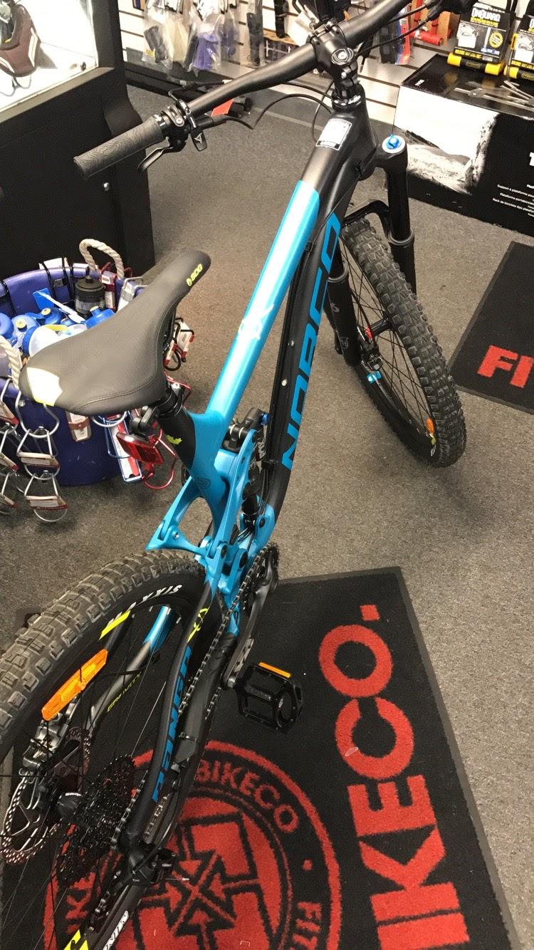 Norco Norco Range Alu 7.2: A close-up view of a blue mountain bike positioned inside a shop, with a focus on its frame, handlebars, and saddle. The bike features thick tires and a modern design, resting on a black mat with a red logo. In the background, various biking accessories and equipment are visible on shelves.