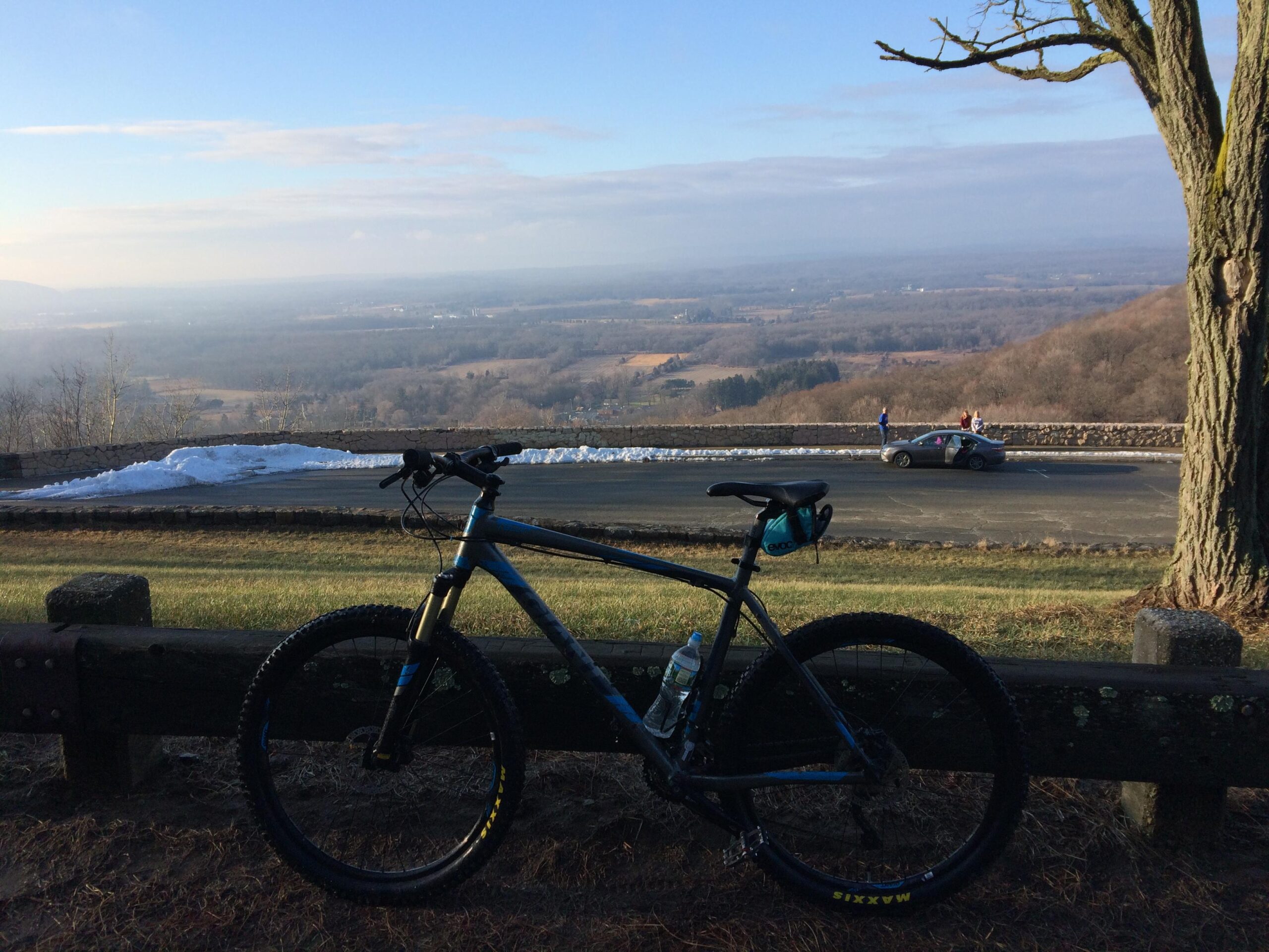 Giant Talon 27.5 2: A mountain bike parked in the foreground with a scenic view of rolling hills and farmland in the background. A small patch of snow is visible on the ground, and a few people stand by a parked car along the road. The sky is mostly clear with soft clouds.