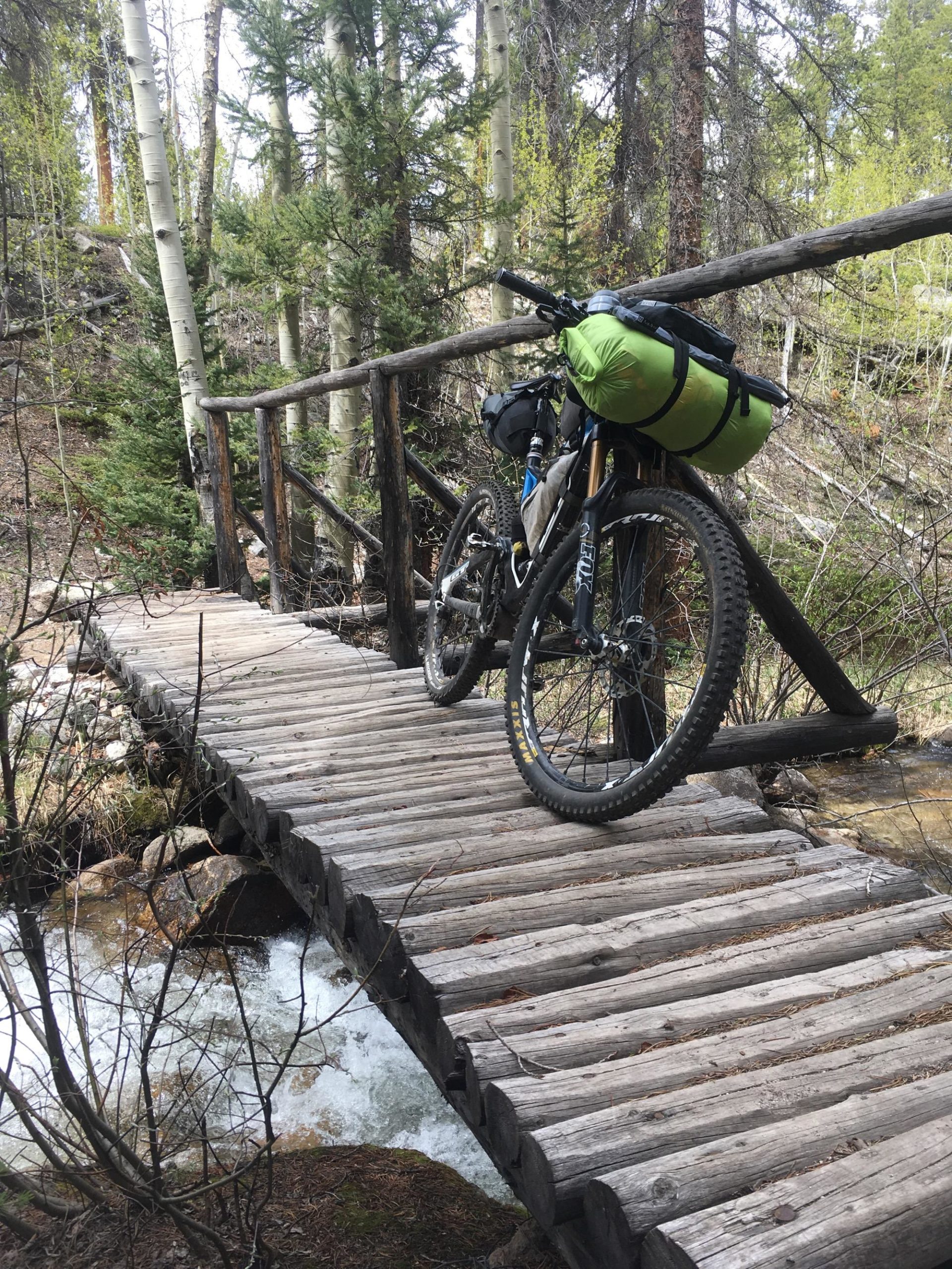 A mountain bike with a green bag attached rests on a rustic wooden bridge over a flowing creek, surrounded by trees and natural vegetation. Colorado Trail: Mt. Shavano thd to Chalk Creek thd mountain bike trail.