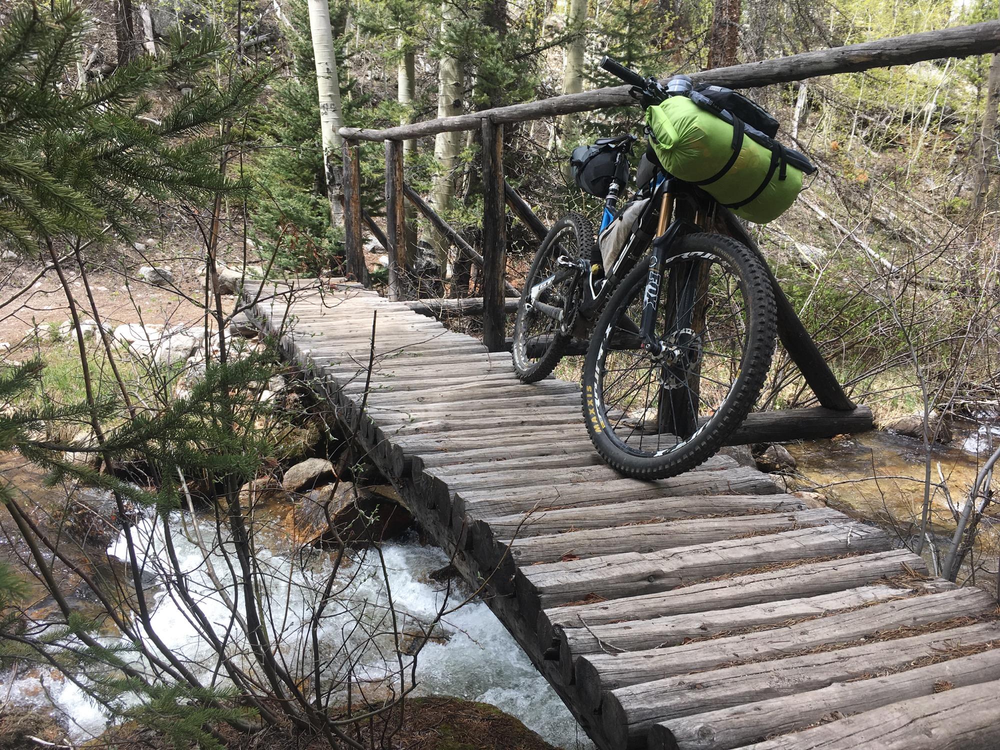 A mountain bike parked on a wooden bridge over a flowing creek, surrounded by trees and greenery in a natural outdoor setting. Colorado Trail: Mt. Shavano thd to Chalk Creek thd mountain bike trail.