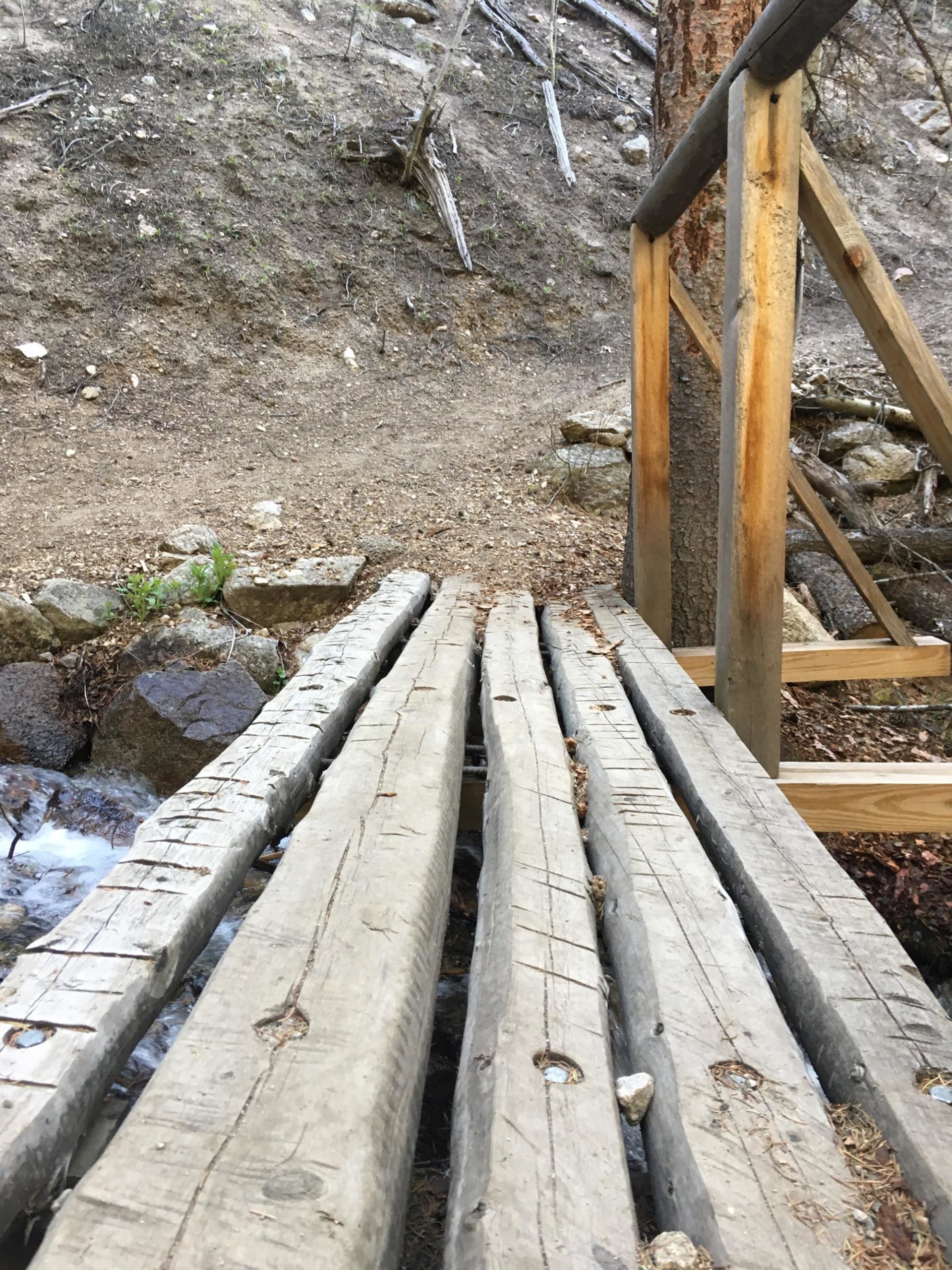 Wooden bridge extending over a small stream, surrounded by rocky terrain and sparse vegetation. The bridge features weathered wooden planks and sturdy railings, leading toward a dirt path on the hillside. Colorado Trail: Mt. Shavano thd to Chalk Creek thd mountain bike trail.