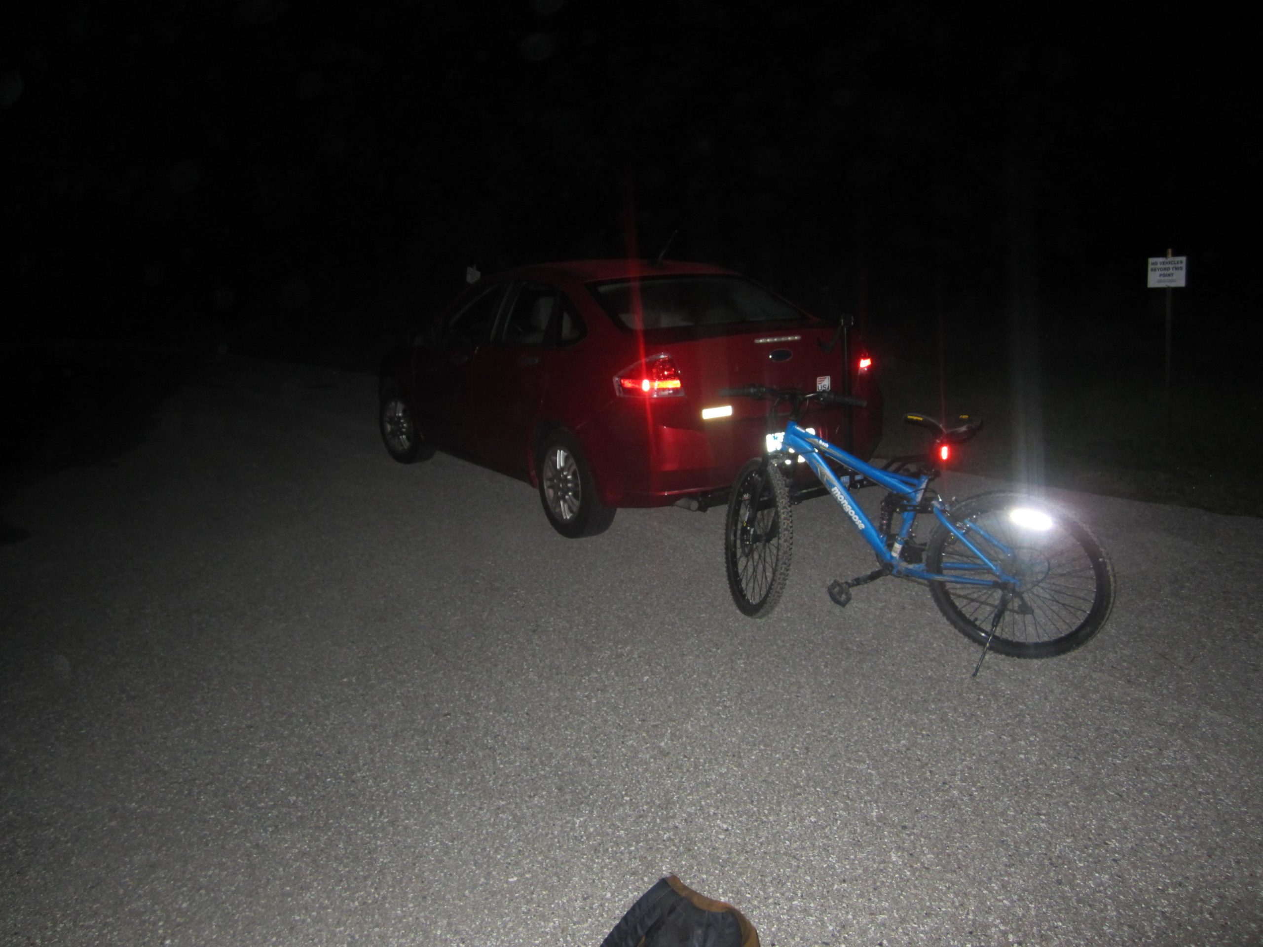 A red car parked on a gravel road at night, with a blue bicycle leaning against it. Reflective elements on the bicycle are visible, and a sign can be seen in the background. The surrounding area is dark, indicating a nighttime setting. Tuckers Grade Out Tram Grade Back mountain bike trail.