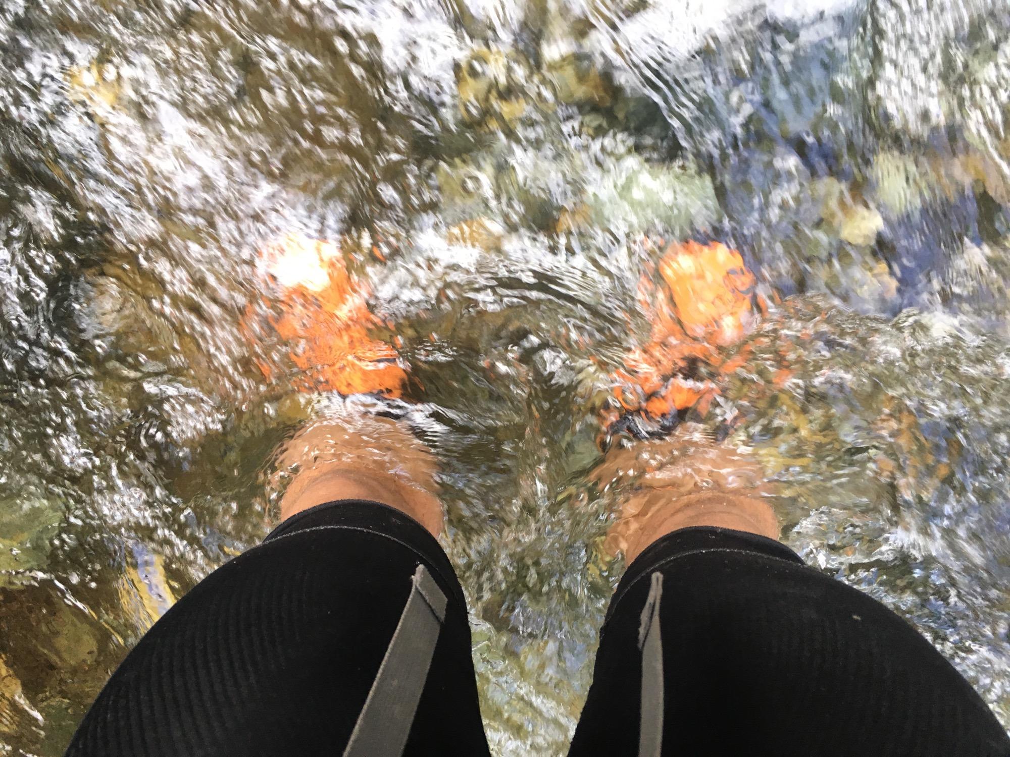 Feet submerged in clear water, with orange water shoes visible. The surrounding water creates ripples and reflects light, and there are rocks and pebbles beneath the surface. The person is wearing dark shorts. Rainbow Trail: Bear Creek Thd to Kerr Gulch Thd mountain bike trail.