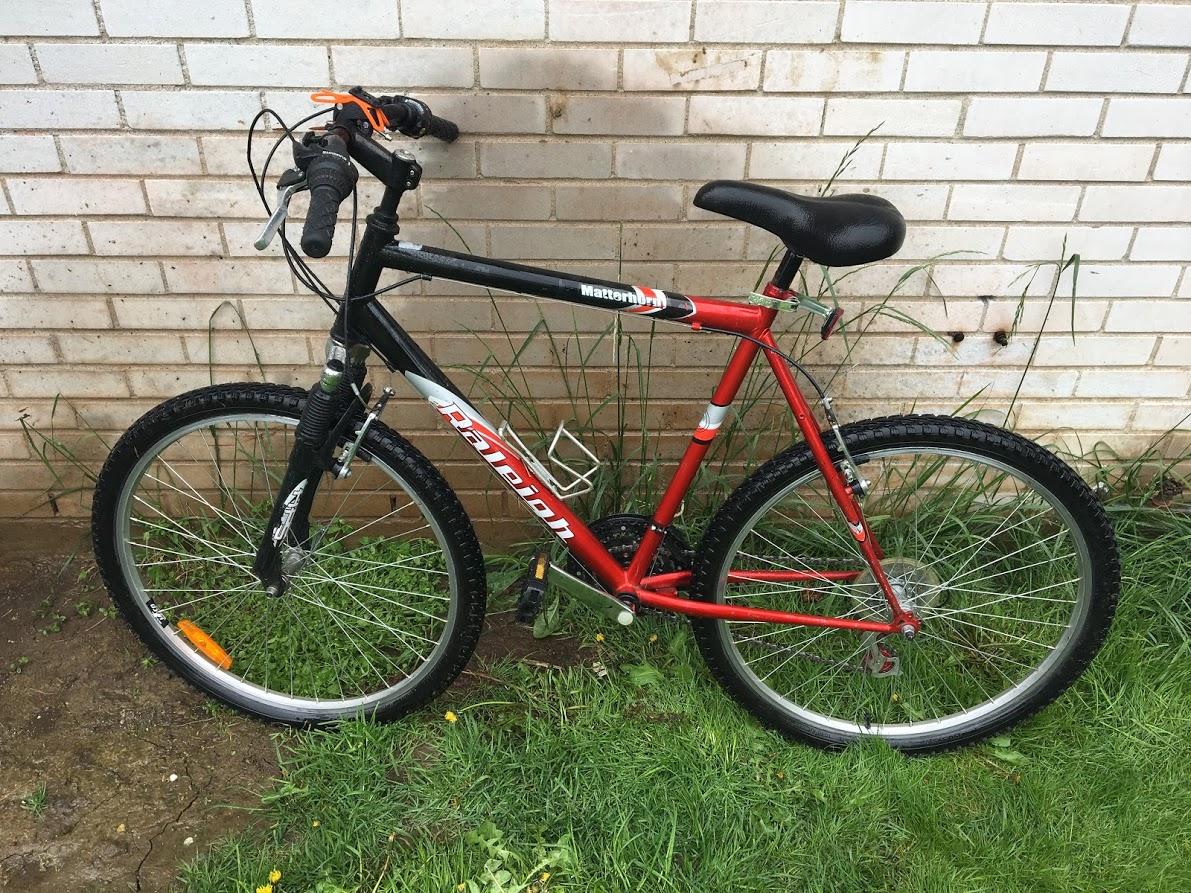 Raleigh Matterhorn: A black and red mountain bike leaning against a white brick wall, surrounded by grass and weeds. The bike features a front suspension, black handlebars with grips, and orange reflectors on the wheels.