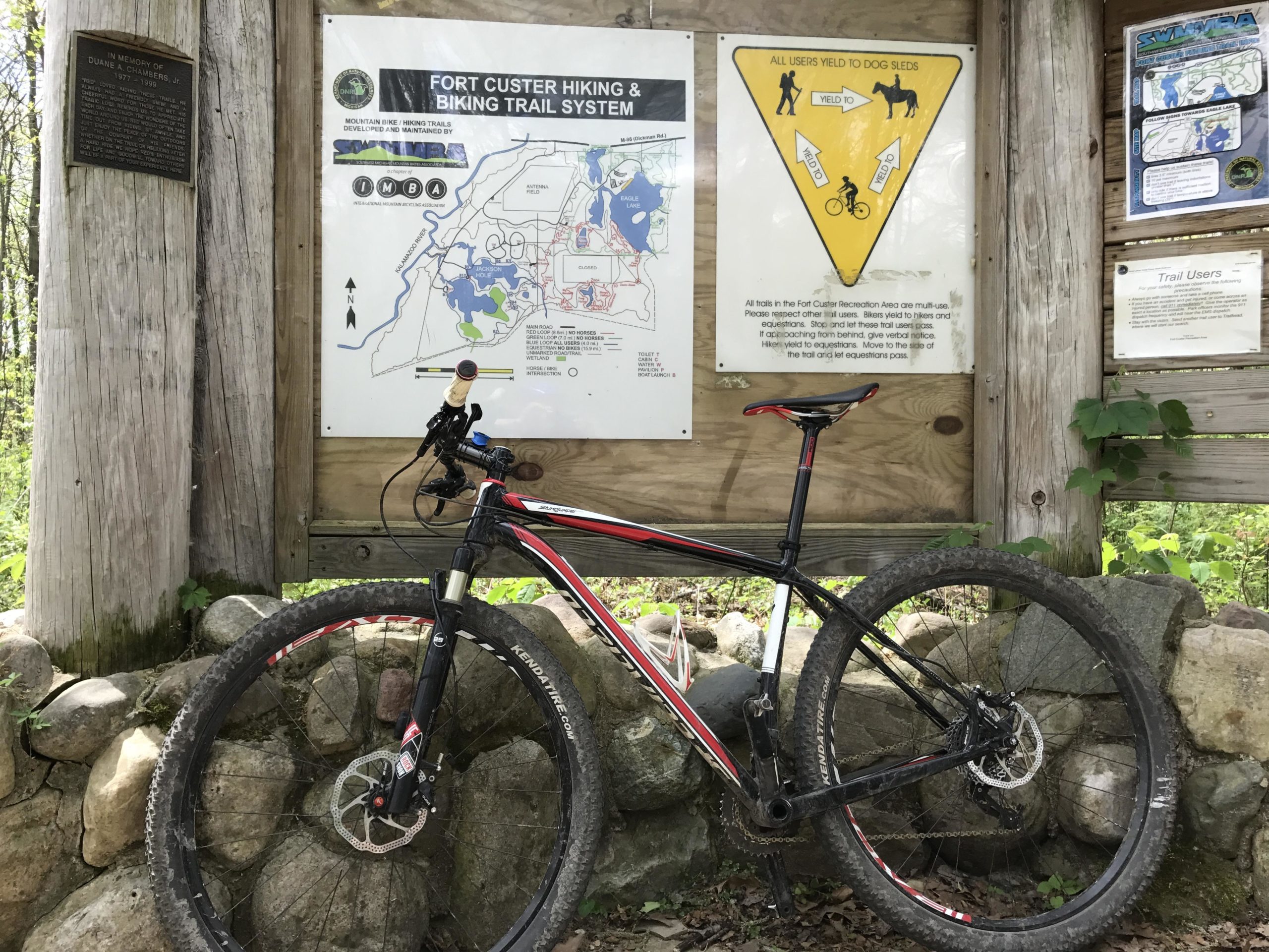 A mountain bike leaning against a stone wall, with a trail map and informational signs posted on a wooden structure in the background. The signs provide details about the Fort Custer hiking and biking trail system, including trail usage guidelines. Lush greenery surrounds the area, indicating a forested environment. Fort Custer Recreation Area mountain bike trail.