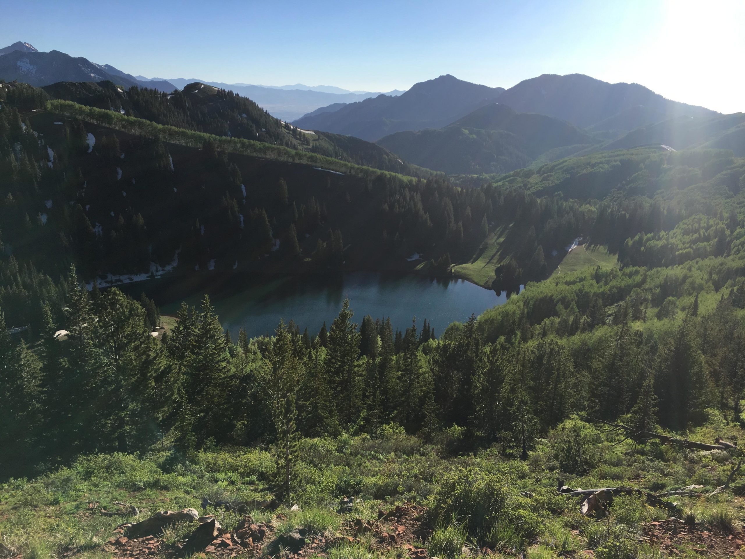 A panoramic view of a serene mountain landscape featuring lush green trees and a tranquil lake nestled between the hills. The scene is bathed in sunlight, showcasing the majestic peaks in the background. Snow can be observed on some areas of the mountains, while the foreground is filled with vibrant foliage and rocky terrain. Scott's Bypass mountain bike trail.