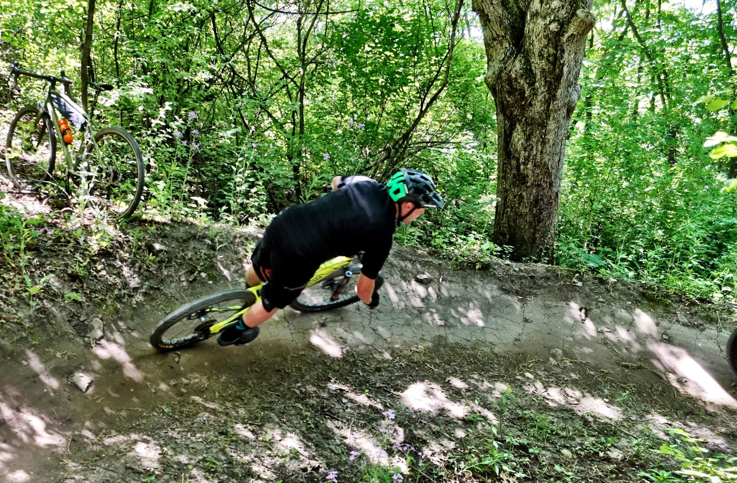 A cyclist wearing a black helmet and jersey leans into a dirt turn on a forest trail, surrounded by lush greenery. A mountain bike is parked nearby, partially obscured by plants. The scene captures the excitement of mountain biking in a natural setting. Quarry Ridge Park mountain bike trail.