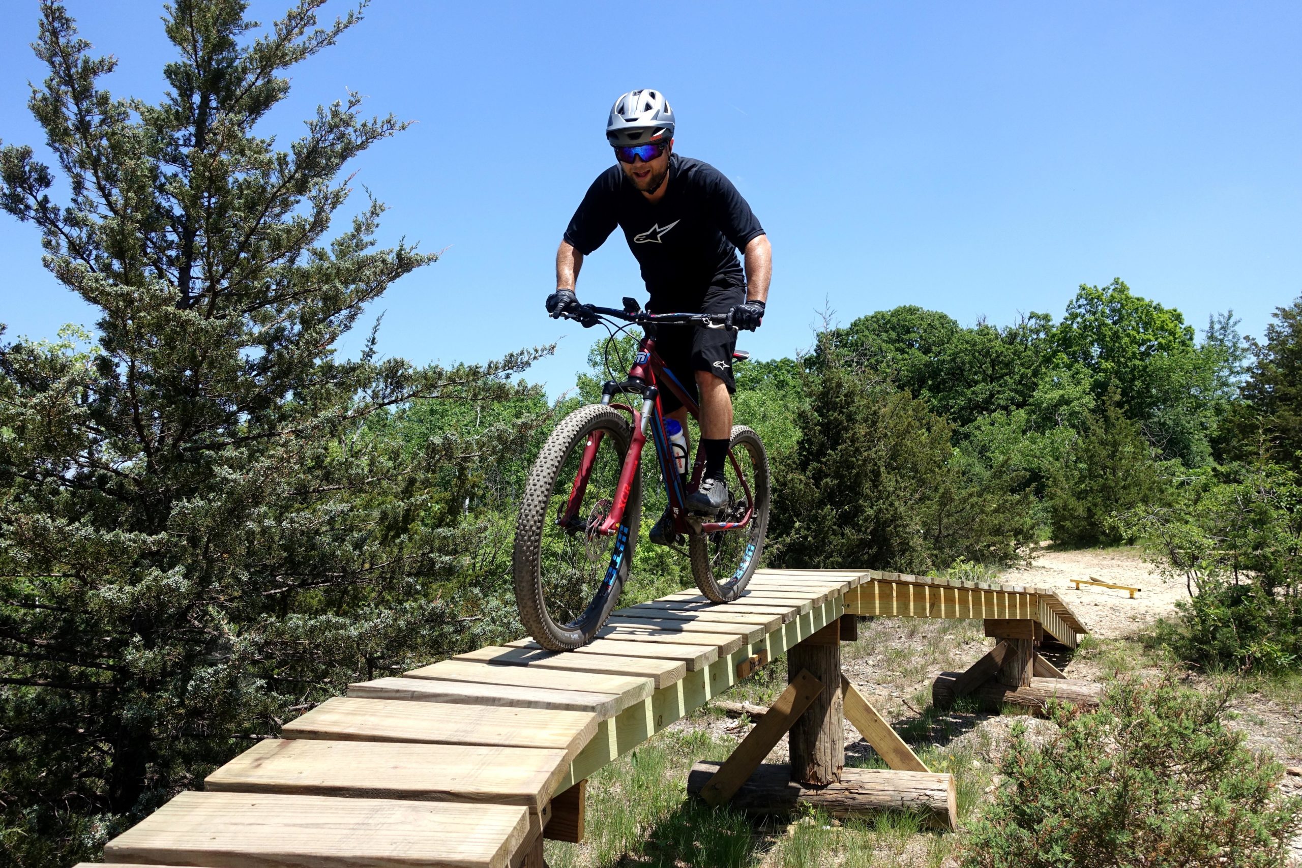 A person riding a mountain bike on a wooden bridge in a natural outdoor setting, surrounded by trees and blue sky. The cyclist is wearing a helmet and sunglasses, appearing focused as they navigate the elevated path. Quarry Ridge Park mountain bike trail.