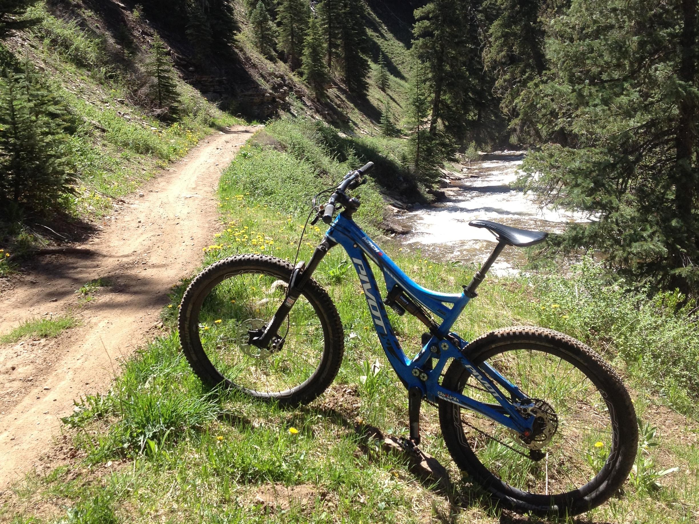 A blue mountain bike is parked beside a winding dirt trail, with a flowing creek visible in the background. The scene is surrounded by lush greenery and tall trees, creating a serene outdoor setting. Hermosa Creek Trail mountain bike trail.