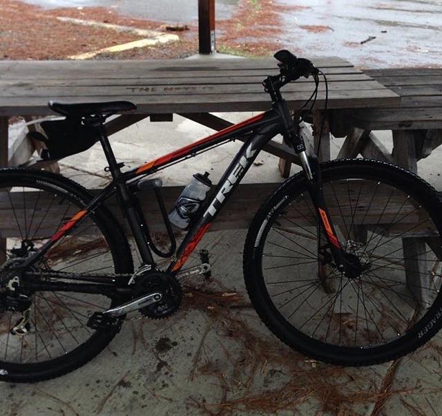 Trek Marlin 5 29er: A black and orange Trek mountain bike is leaning against a picnic table. A water bottle is attached to the frame, and the ground is wet, indicating recent rain. The background shows slatted wooden benches and a blurred outdoor setting.