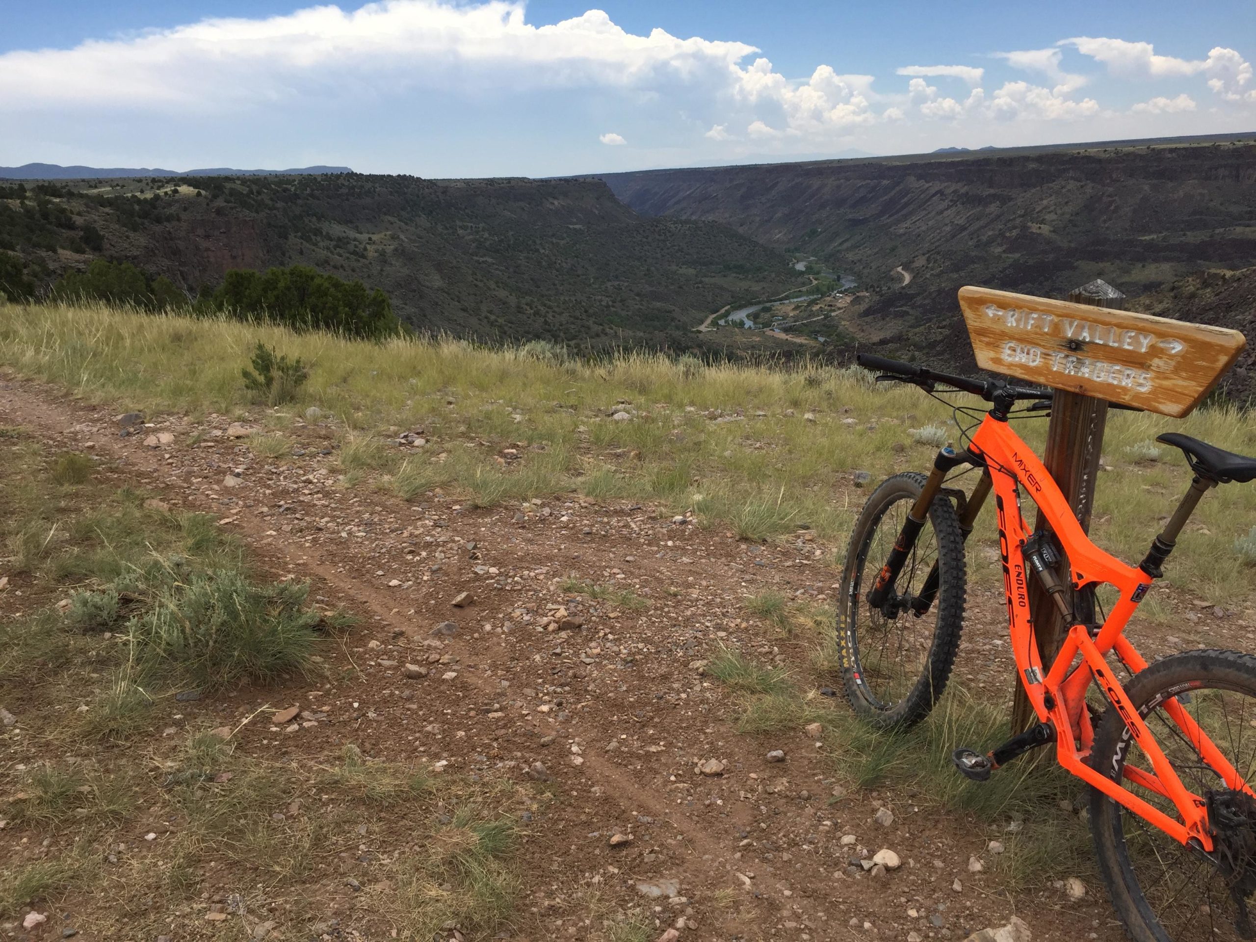 A vibrant orange mountain bike is leaning against a wooden signpost that reads "Rift Valley End Trail" in a scenic outdoor landscape. In the background, a vast canyon with green vegetation stretches out under a partly cloudy sky. A dirt trail with rocks and grass is visible in the foreground, leading towards the canyon view. Taos Valley Overlook mountain bike trail.