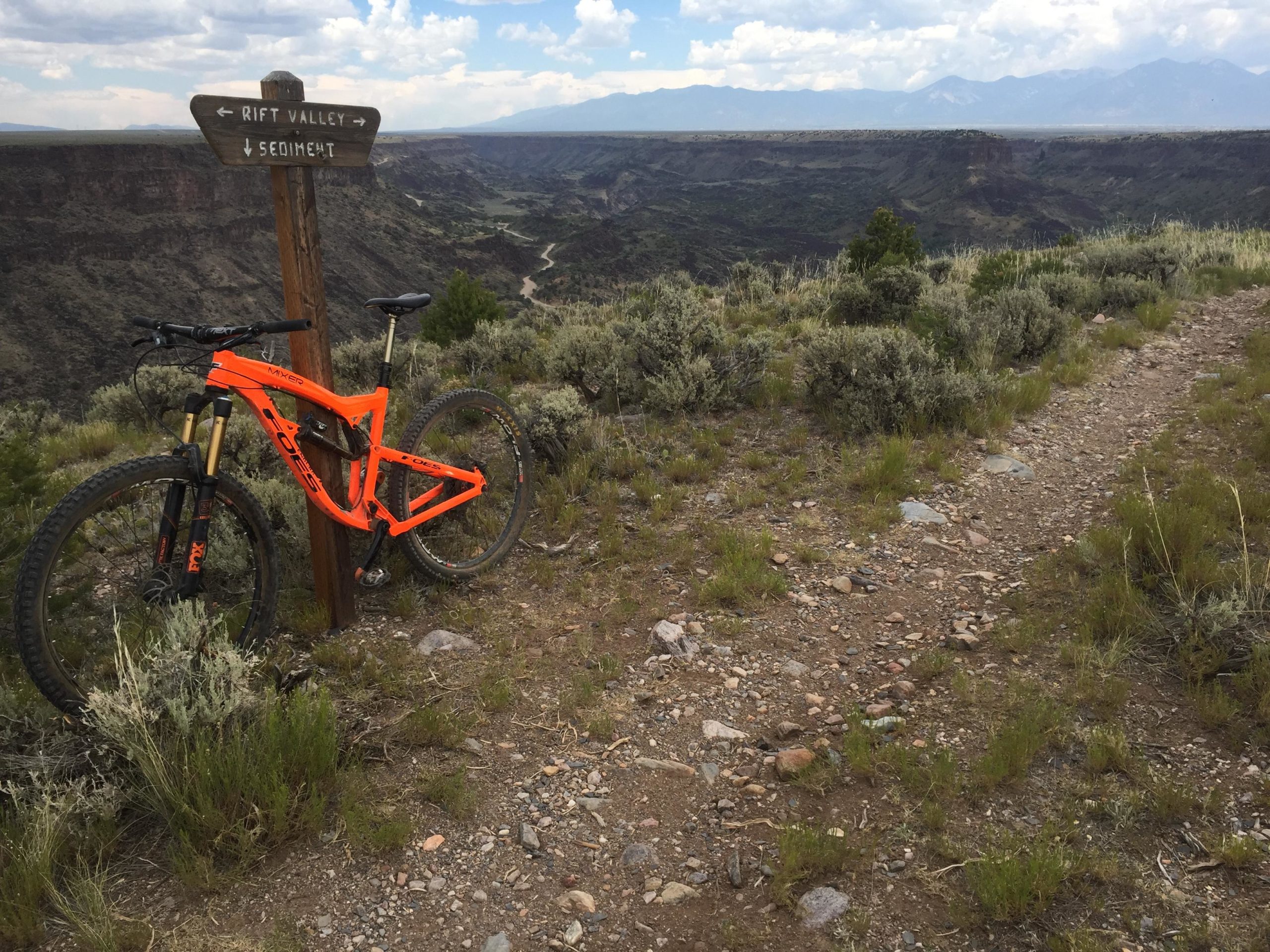 Mountain bike parked next to a wooden trail sign marked "Rift Valley" and "Sediment," overlooking a canyon landscape with sparse vegetation and mountains in the background under a partly cloudy sky. Taos Valley Overlook mountain bike trail.
