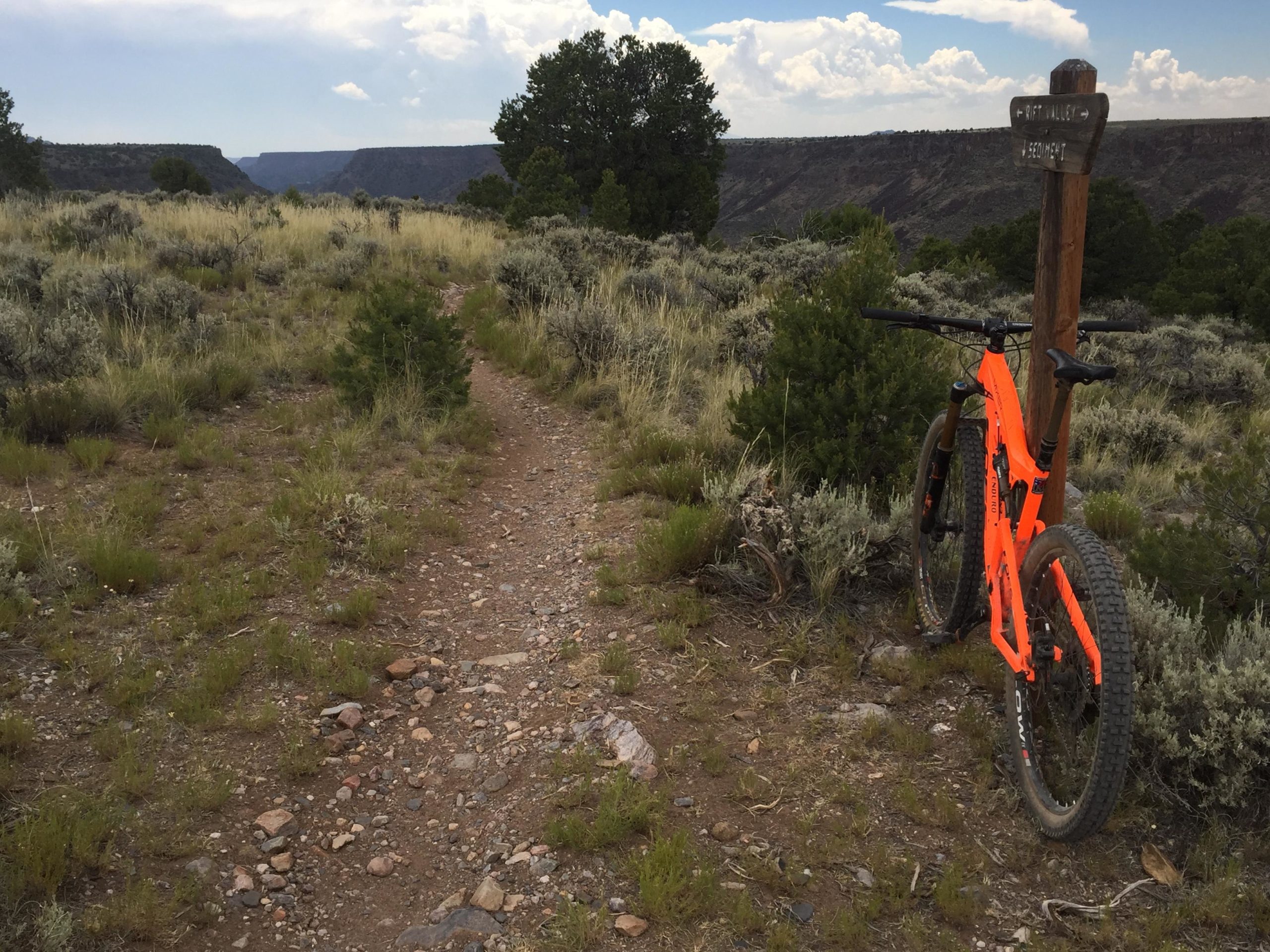 A vibrant orange mountain bike rests against a wooden trail sign in a rugged outdoor landscape. The dirt path, flanked by light green shrubbery and rocky terrain, meanders towards a distant canyon under a partly cloudy sky. Taos Valley Overlook mountain bike trail.