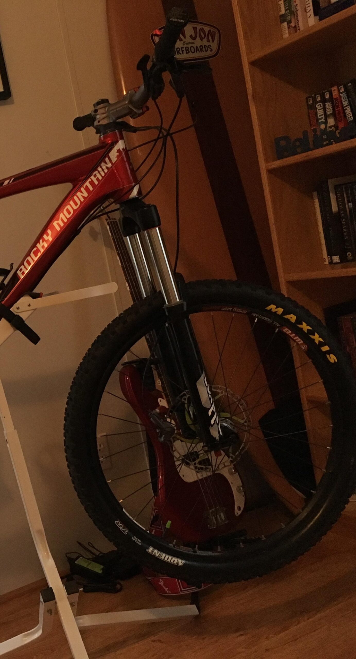 A red Rocky Mountain mountain bike is propped up on a bike stand in a cozy indoor setting. In the background, a surfboard is partially visible alongside a bookshelf filled with books and a red guitar. The bike features black Maxxis tires and a silver front suspension fork.