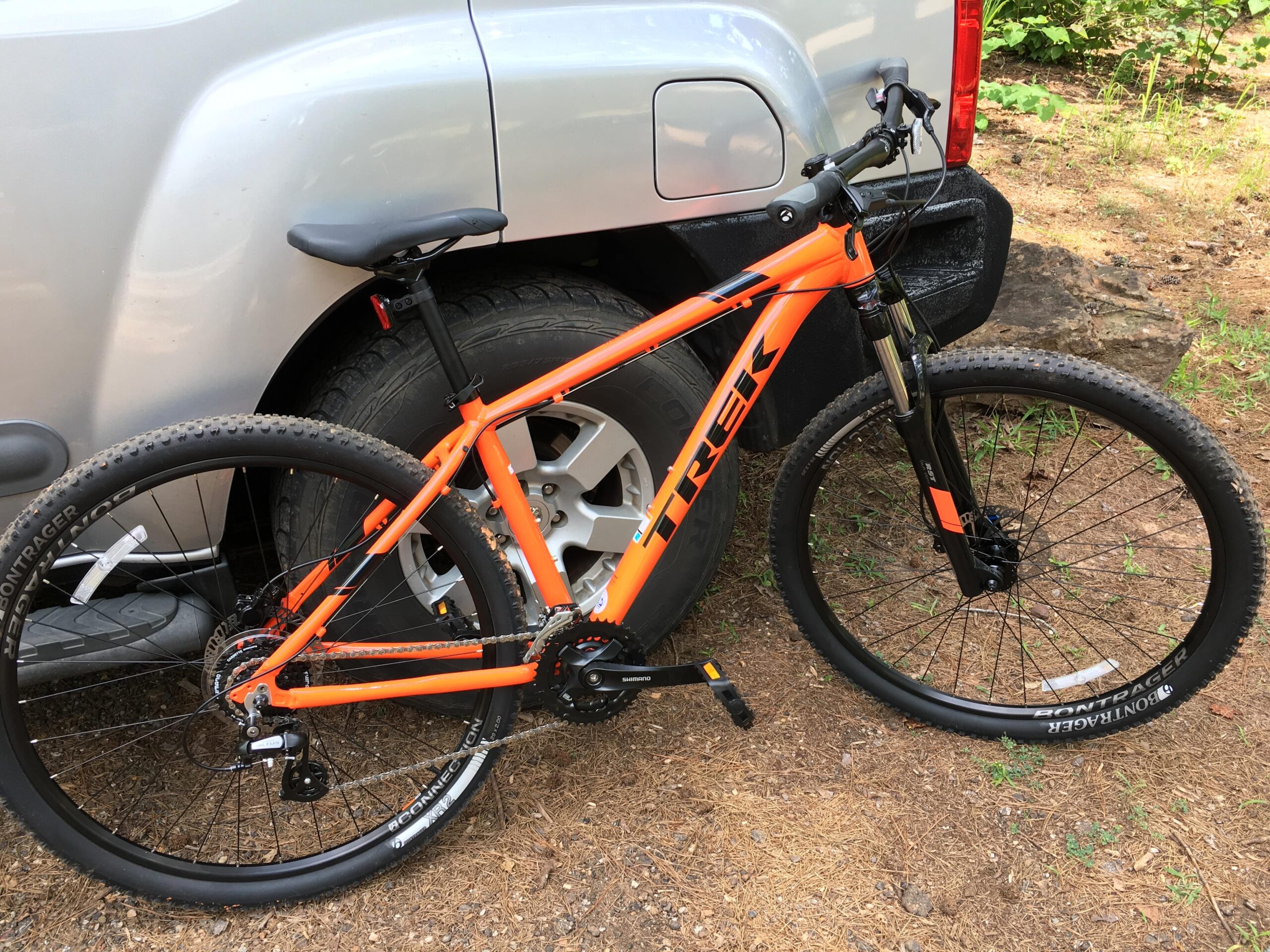 Trek Marin 6: A bright orange Trek mountain bike parked next to a silver vehicle, with its wheels resting on a dirt surface surrounded by greenery.