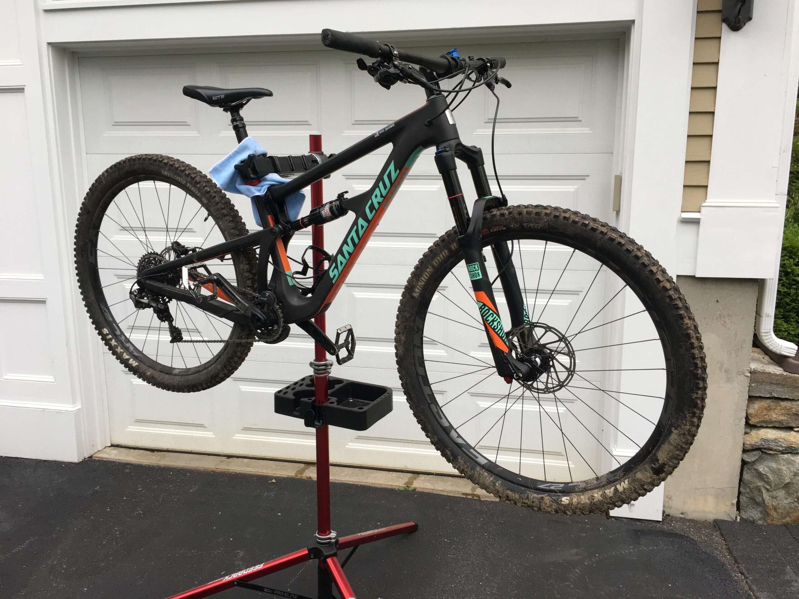 Santa Cruz Hightower 29er/27.5: Mountain bike elevated on a repair stand, showcasing its black frame with colorful accents. The bike has a dirty rear wheel and is positioned in front of a garage door. A blue cloth is draped over the saddle for maintenance purposes.