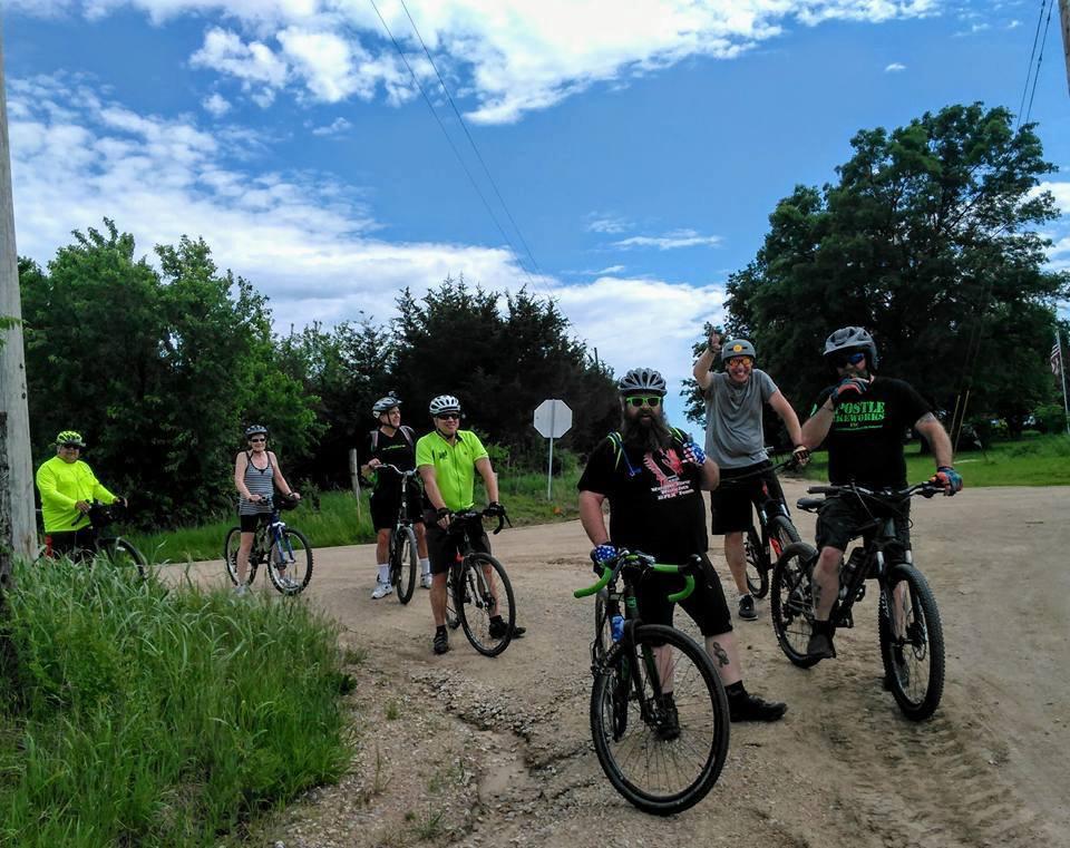 Specialized Rockhopper 29: A group of seven people, wearing helmets and various cycling attire, are gathered on a dirt road. They are on mountain bikes and pose for the camera, smiling and showing a sense of camaraderie. The background features greenery and a partly cloudy sky.