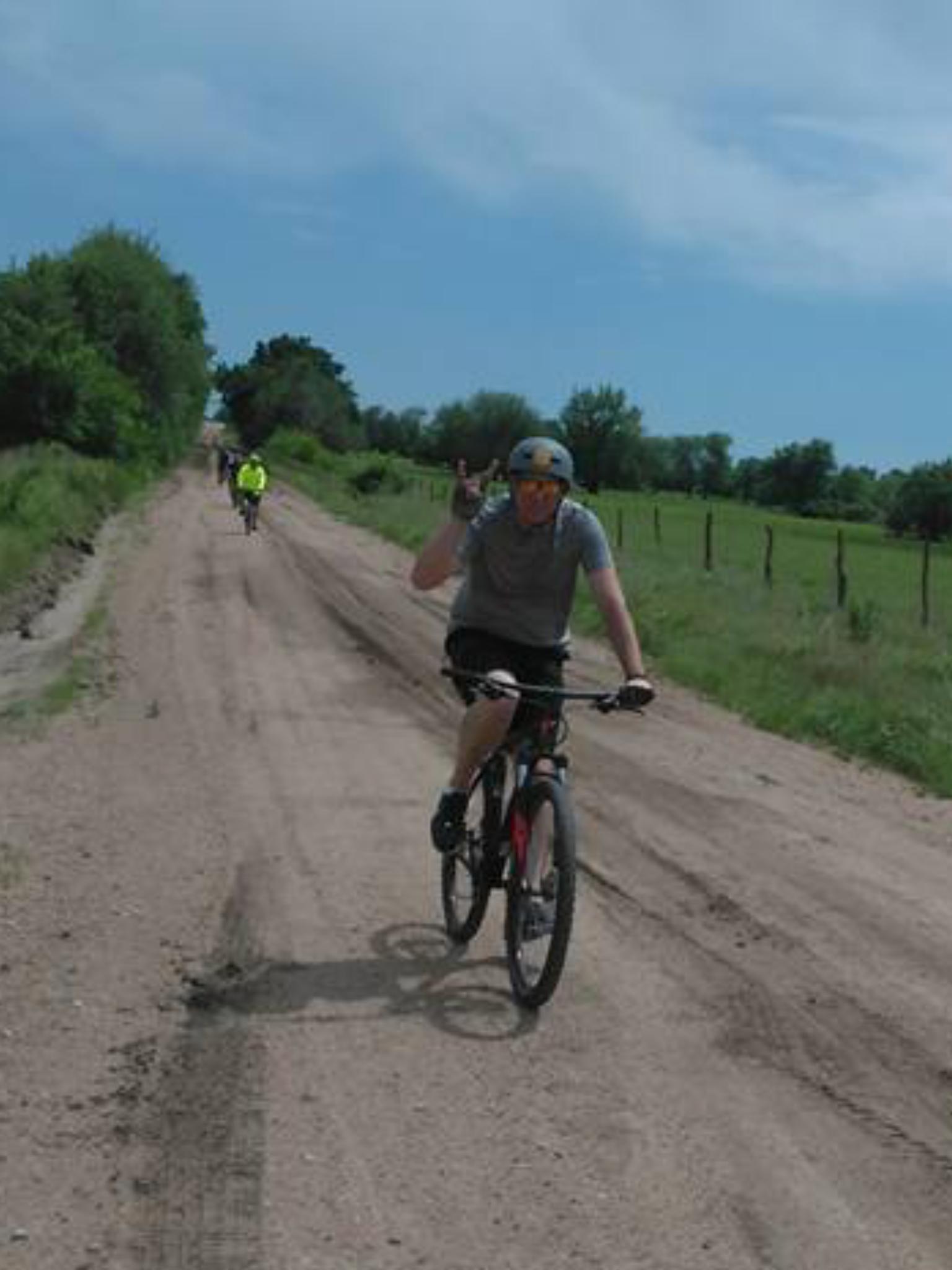 Specialized Rockhopper 29: A person riding a mountain bike on a dirt road, wearing a helmet and a gray shirt, giving a peace sign with one hand. In the background, another cyclist in a reflective yellow jacket can be seen on the same path, surrounded by greenery and under a blue sky.
