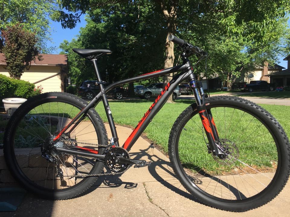 Specialized Rockhopper 29: A black and red mountain bike parked on a paved driveway, surrounded by grass and trees. The bike features thick tires and a sturdy frame, positioned in a residential area with houses and parked cars visible in the background.