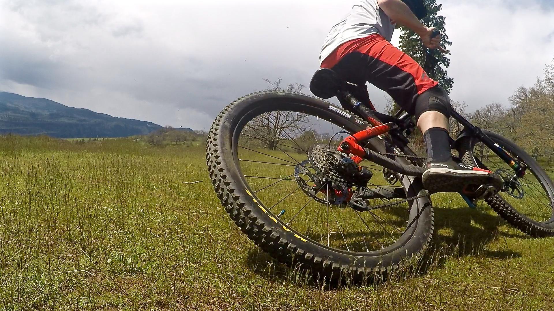Maxxis Rekon: A mountain biker making a sharp turn on a grassy landscape, with a focus on the bike's rear tire and rider's legs. The background features a scenic view of rolling hills under a cloudy sky.