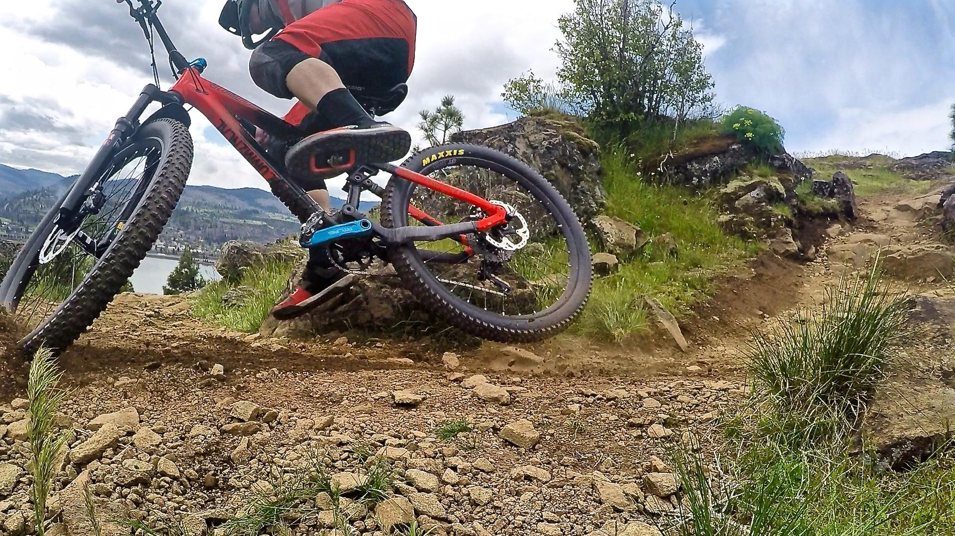 Maxxis Rekon: A mountain biker in red shorts and a helmet navigates a rocky trail, leaning into a turn on a red and black bicycle. Lush greenery and a scenic landscape are visible in the background under a partly cloudy sky.