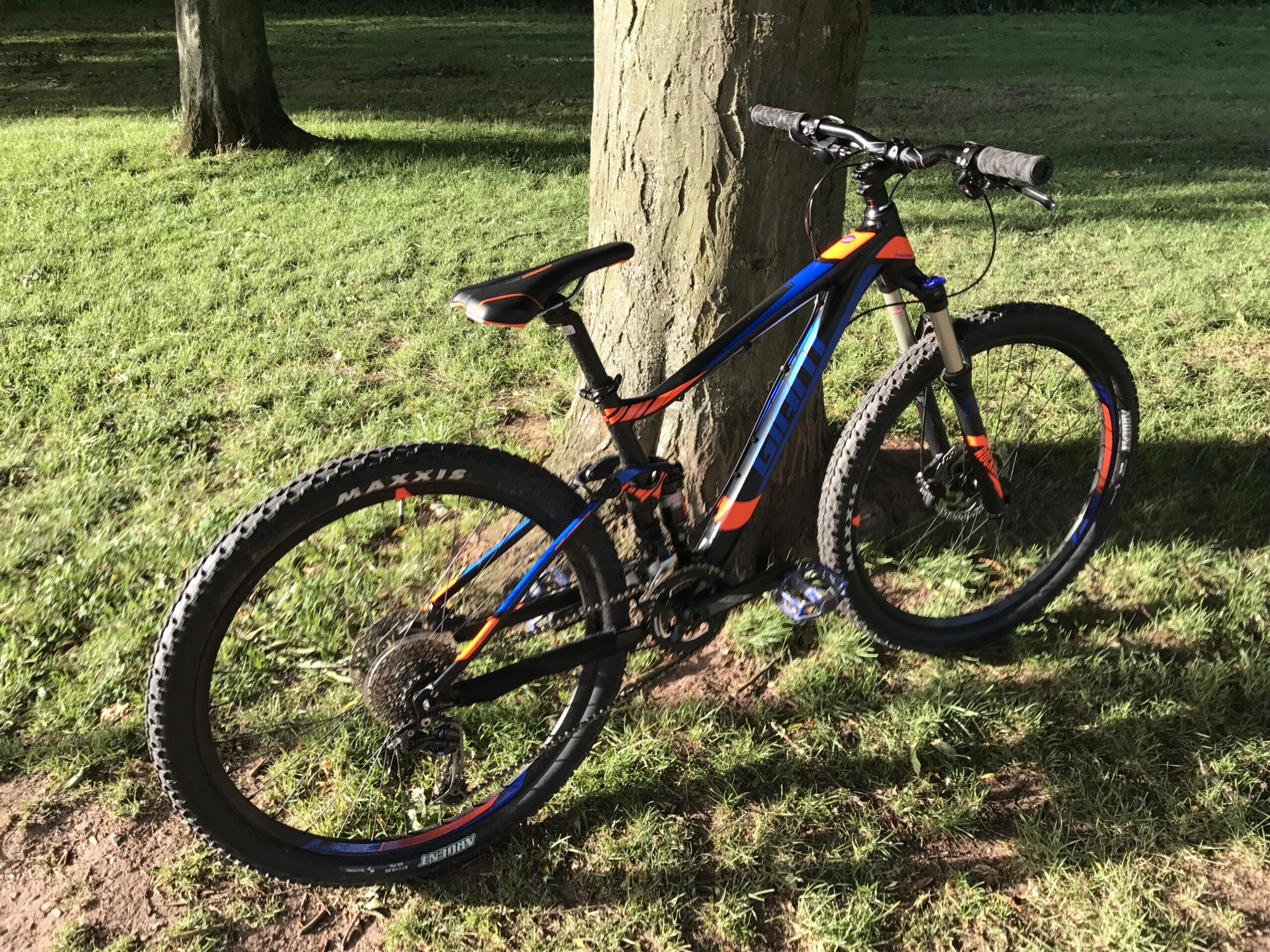 Giant Stance 27.5 2: A colorful mountain bike leaning against a tree, positioned on grassy ground with sunlight casting shadows. The bike features black and orange accents, along with wide tires designed for off-road terrain.
