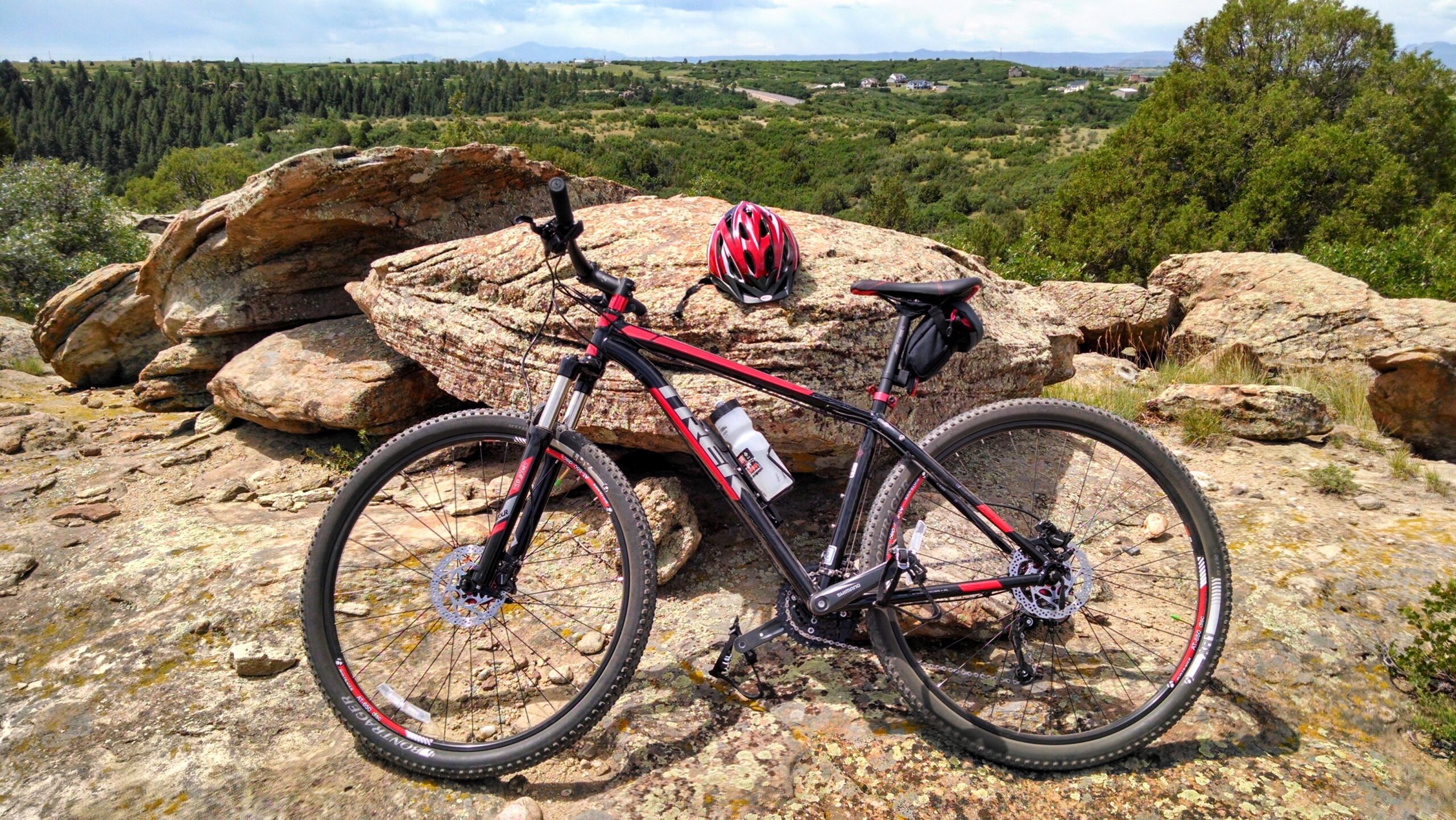 Trek Excalibur: A black and red mountain bike is parked on rocky terrain, with a helmet positioned on a large rock nearby. Lush green hills and trees are visible in the background under a partly cloudy sky. A water bottle is attached to the bike’s frame.
