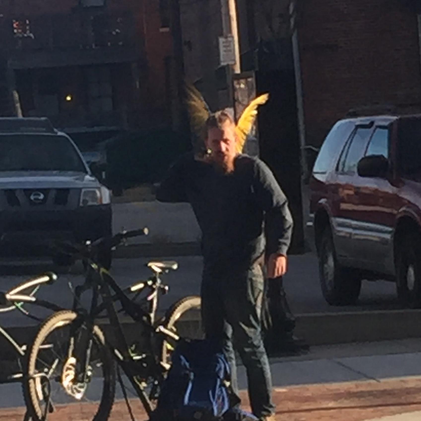Specialized Stumpjumper FSR: A person with a beard and long hair, wearing a dark sweater and jeans, standing outdoors next to a bicycle. They are holding their hand to their ear, suggesting they are on a call. The background features parked vehicles and a city setting. Bright yellow feathers are visible in their hair, adding a whimsical touch to their appearance.