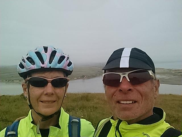 A selfie of two cyclists on a scenic overlook, wearing helmets and sunglasses, with a foggy landscape in the background featuring water and green hills. Both are dressed in bright yellow jackets for visibility. Estero Trail To Sunset Beach mountain bike trail.