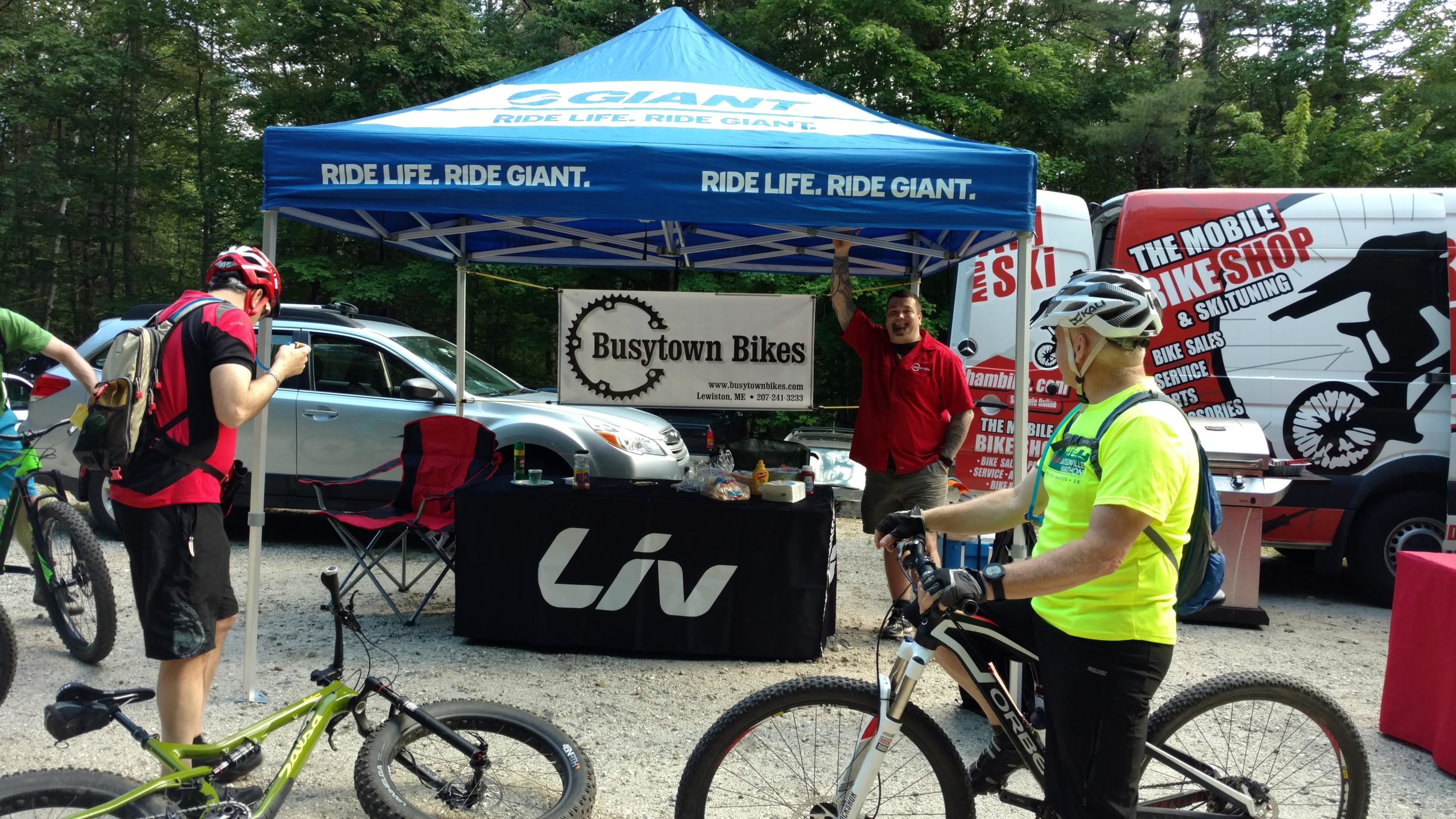 A vibrant outdoor setup featuring a blue canopy promoting "Giant" bicycles, with a sign for "Busytown Bikes" visible. Two cyclists are present: one in a red helmet and shirt checking a device, and another in a bright yellow shirt examining his bike. A parked gray vehicle and a colorful mobile bike shop are in the background, surrounded by lush greenery. Bradbury Mt State Park mountain bike trail.