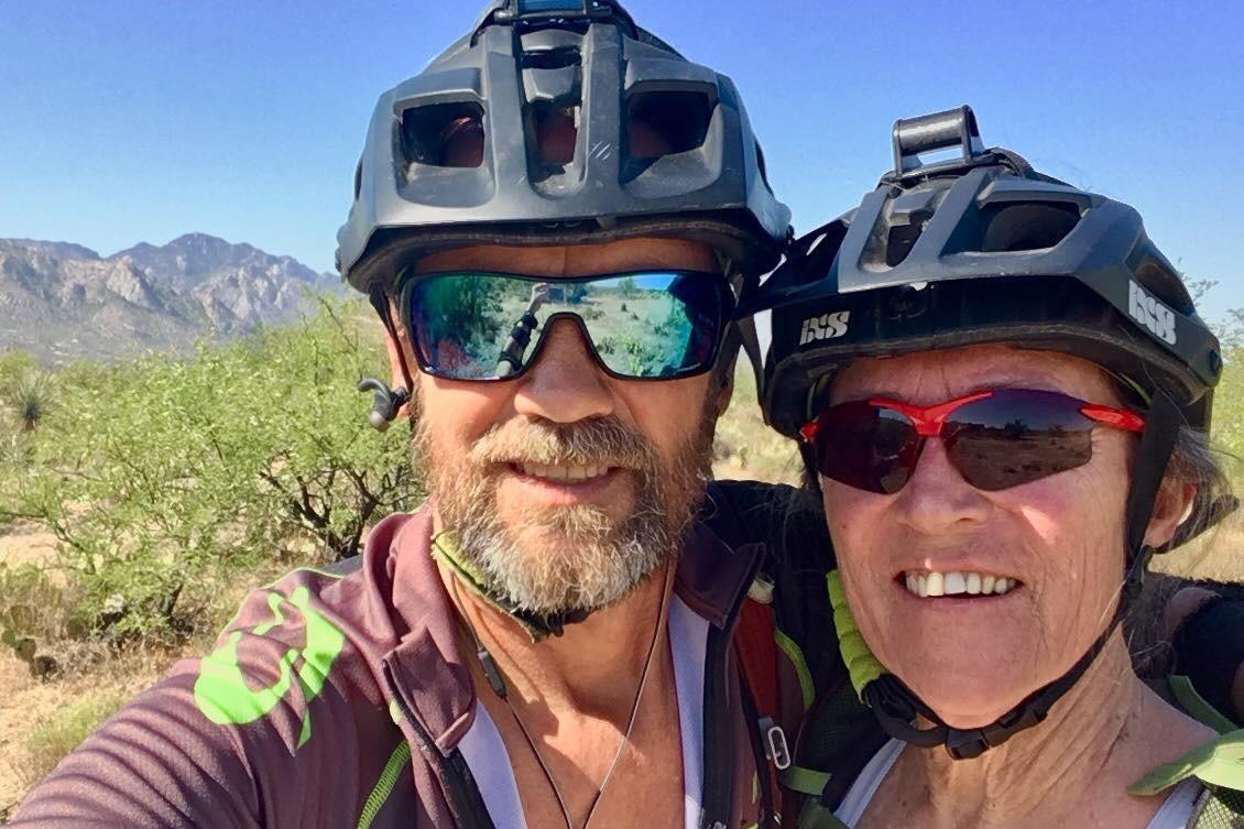 Alt tag: A smiling couple wearing mountain biking helmets and sunglasses poses for a selfie outdoors, with a landscape of mountains and vegetation in the background. 50-year Trail / Golder Ranch mountain bike trail.
