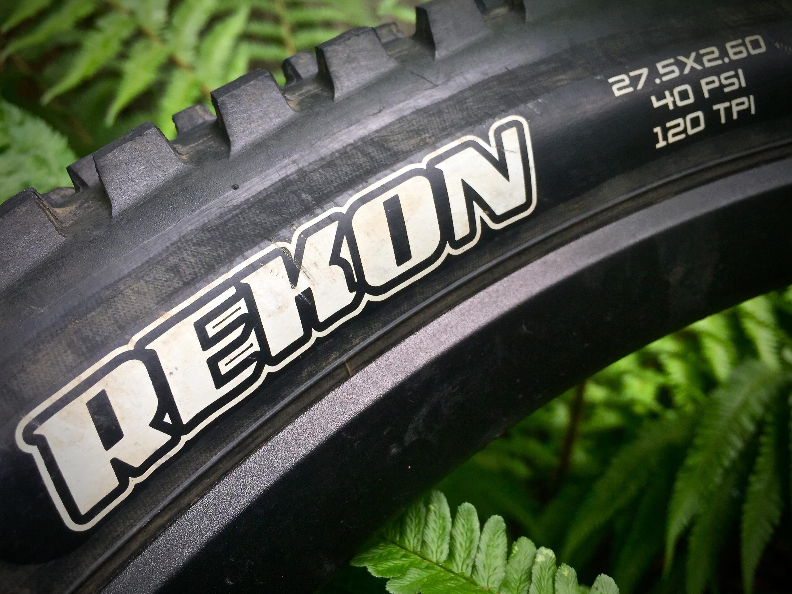 Maxxis Rekon: Close-up of a mountain bike tire showcasing the "Rekon" brand logo, with visible specifications including tire size (27.5 x 2.60), recommended pressure (40 PSI), and thread count (120 TPI), set against a backdrop of green ferns.