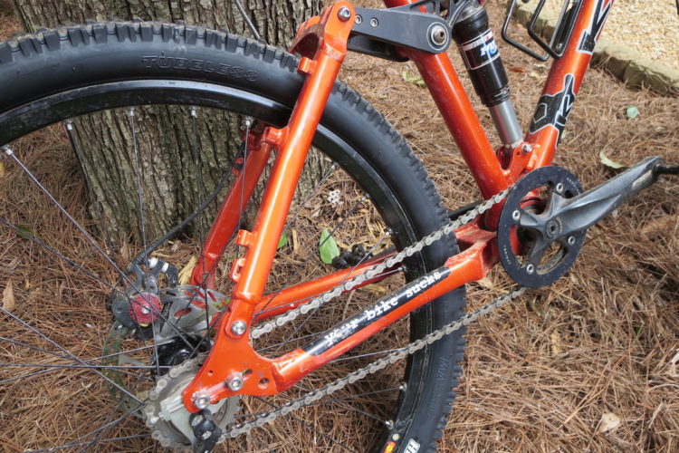 A close-up view of the rear suspension and drivetrain of an orange mountain bike, showcasing the intricate parts including the rear tire, chain, and shock absorber. The bike is positioned near a tree, with pine needles scattered beneath it.