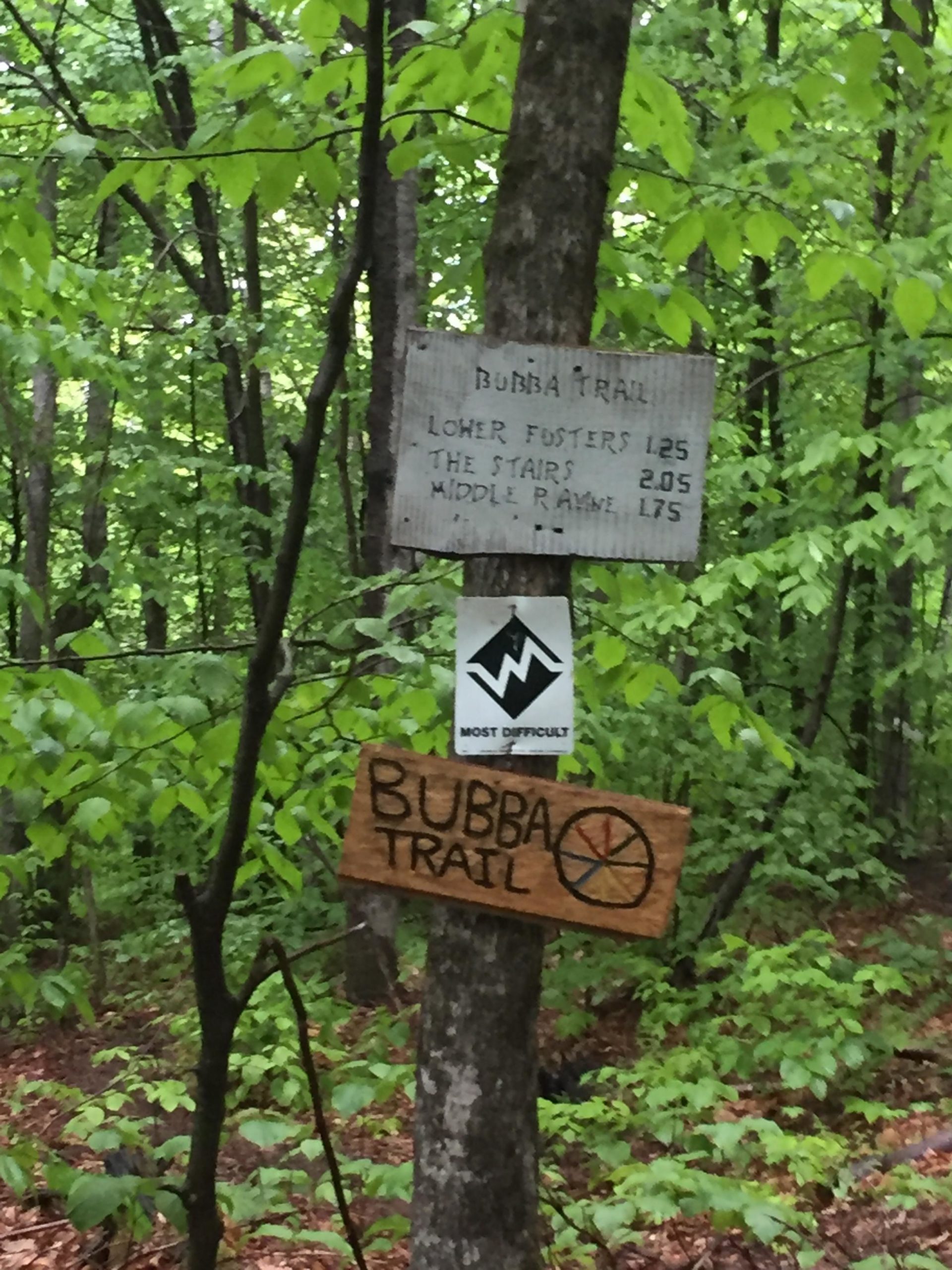 Wooden trail signs indicating "Bubba Trail" and its various routes, surrounded by lush green trees and foliage. The upper sign lists trail options with their respective distances, while a lower sign marks "BUBBA TRAIL" with a mountain biking symbol, indicating the trail's difficulty level as "most difficult." Green Mountain Trails mountain bike trail.