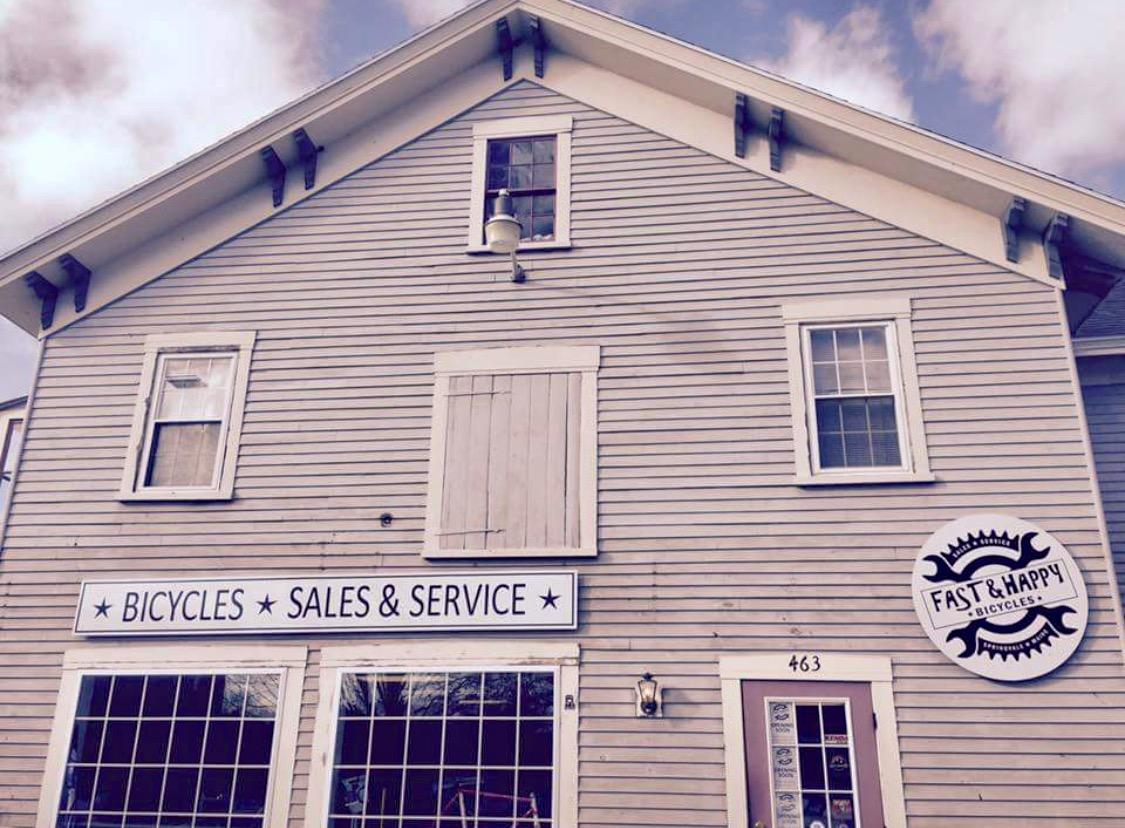 Image of a two-story building with a light gray wooden exterior. The front features a large sign that reads "BICYCLES SALES & SERVICE" and a circular logo for "FAST & HAPPY BICYCLES." Several windows are visible, with one partially obscured by a wooden cover. The building is set against a mostly cloudy sky.