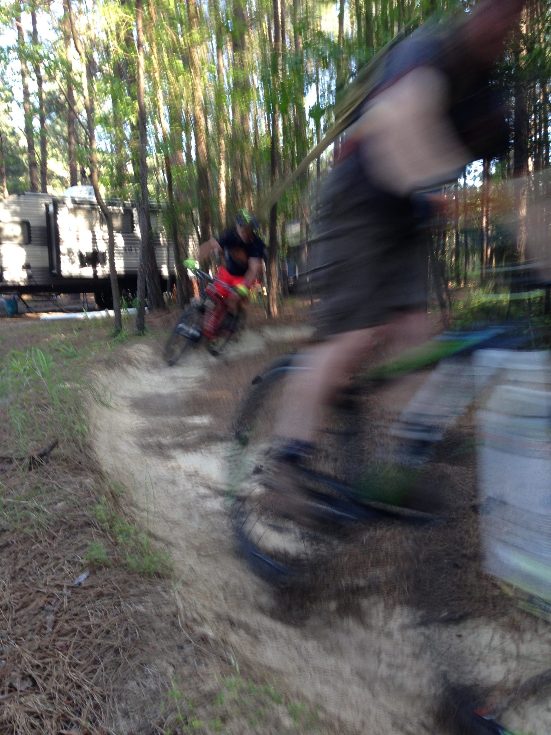 Two mountain bikers racing through a wooded trail, with trees in the background. The image captures the motion of the riders, creating a sense of speed, as they navigate a curved path. A recreational vehicle is partially visible in the background, indicating an outdoor camping scene. Mt. Zion Bike Trails mountain bike trail.