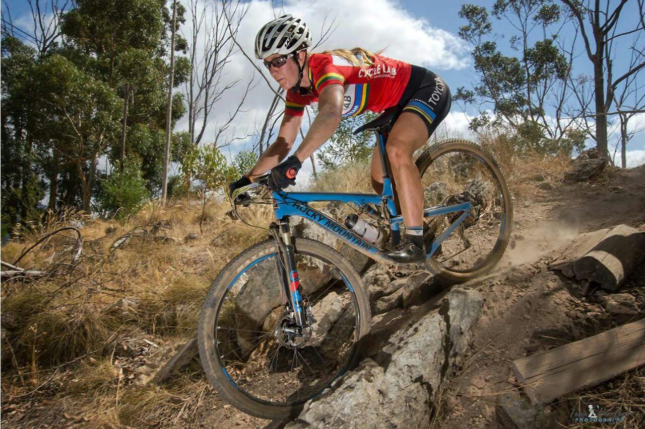 Rocky Mountain Element 970 RSL: A mountain biker navigates a rocky trail, kicking up dust as she rides a blue Rocky Mountain bike. She is wearing a bright red jersey with multi-colored stripes and black shorts, with a helmet and sunglasses for safety. The background features tall trees and a partly cloudy sky.