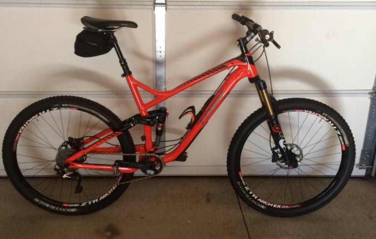 Trek Remedy 9 27.5/650b: A bright orange mountain bike with black and silver accents, featuring a dual suspension frame, wide tires, and a small black saddle bag attached to the seat. The bike is positioned against a garage door background.