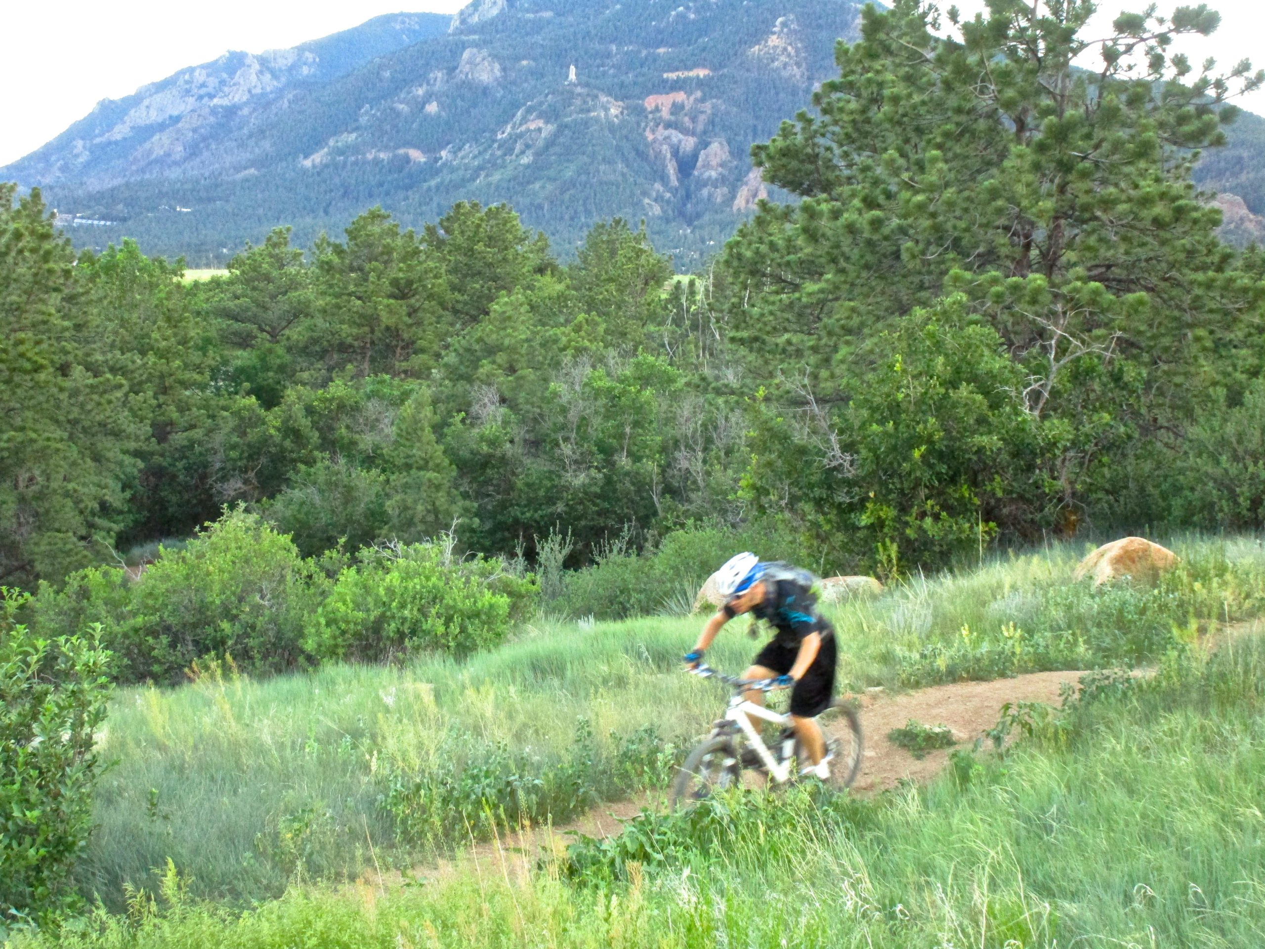 A mountain biker riding on a dirt trail surrounded by lush greenery, with mountains in the background. Stratton Open Space / The Chutes mountain bike trail.