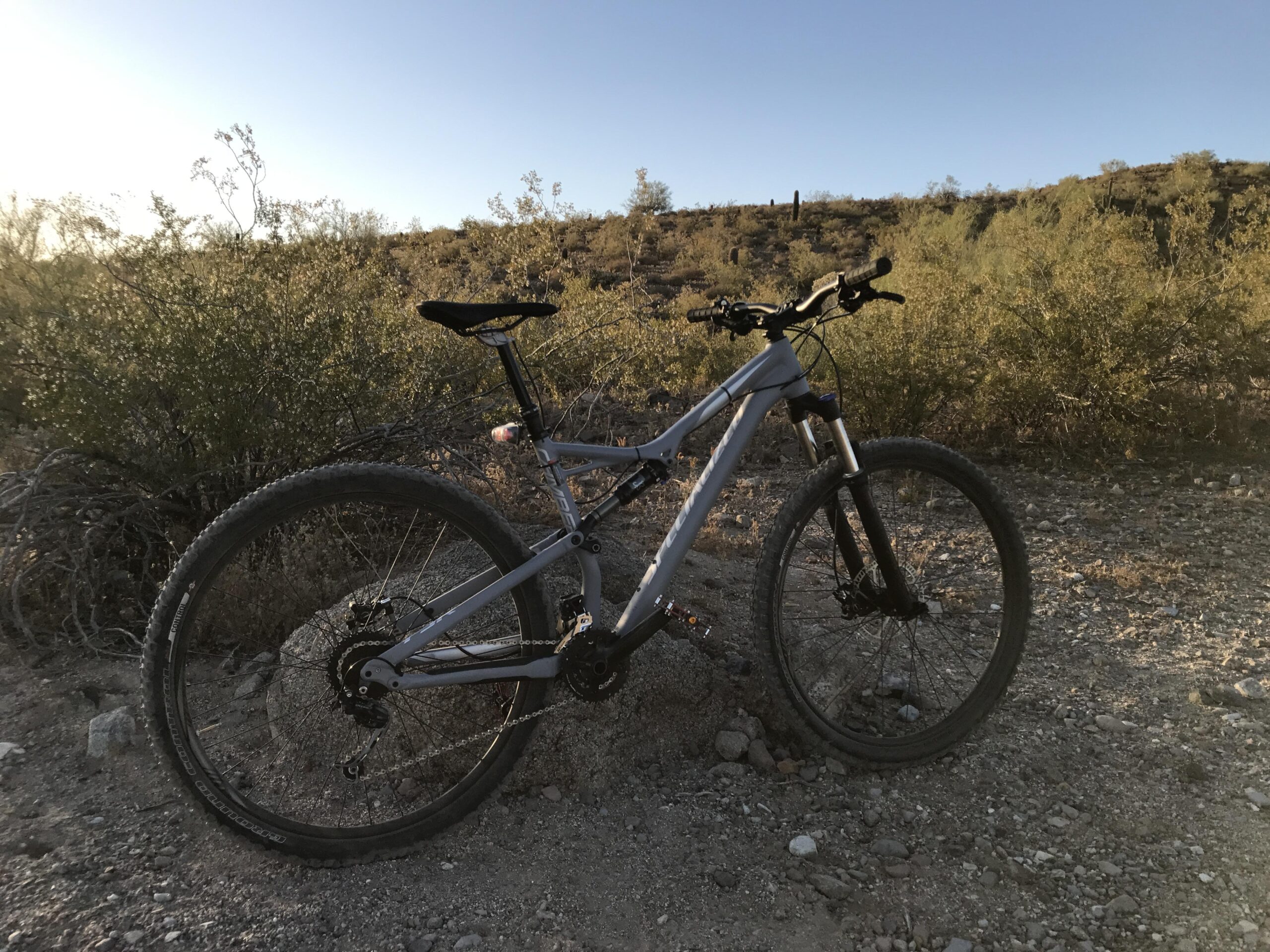 Specialized Camber Comp 29: A mountain bike parked on a dirt trail surrounded by desert vegetation and hills under a clear blue sky. The sun is low on the horizon, casting a warm light on the scene.
