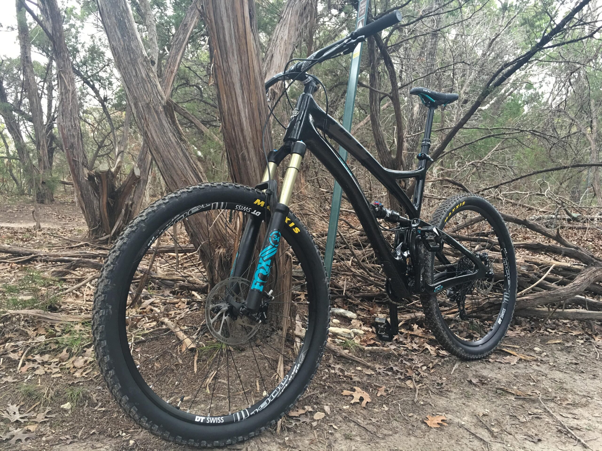 Yeti SB5 Enduro: A black mountain bike leaned against a wooden post, surrounded by trees and underbrush in a natural outdoor setting. The bike features thick tires and a suspension fork, indicating it is designed for rugged terrain.