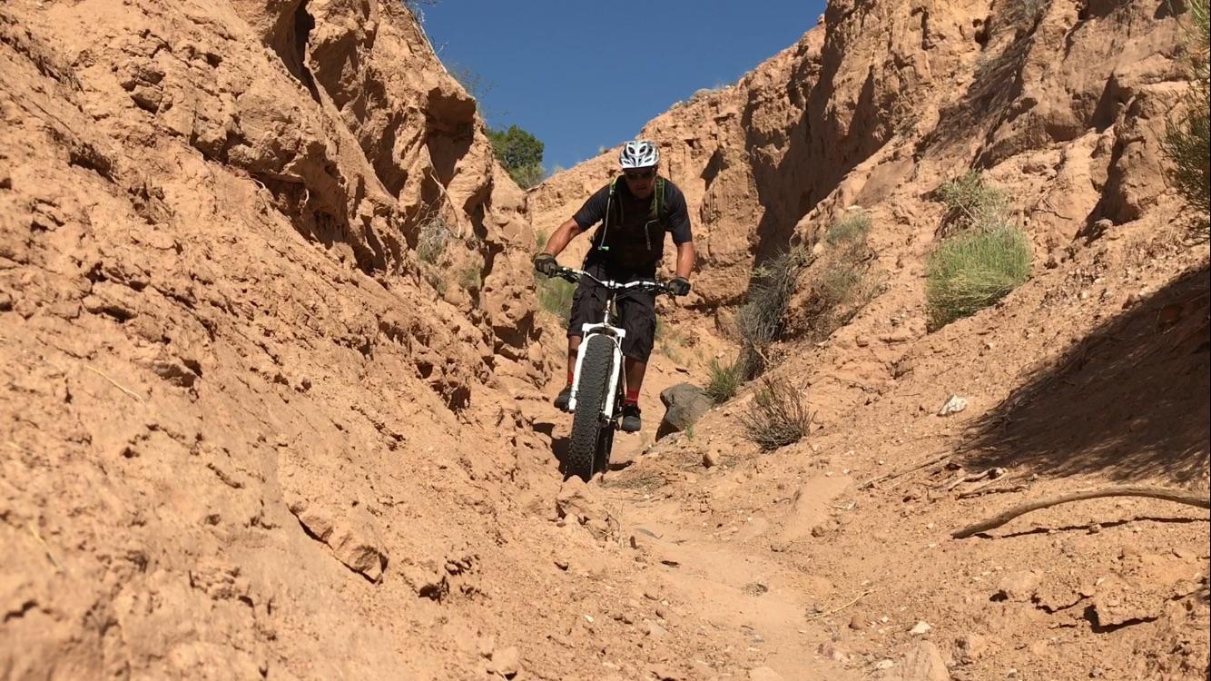 A cyclist riding a fat-tire mountain bike through a narrow, rocky canyon trail surrounded by dry, sandy terrain and sparse vegetation under a clear blue sky. Mariposa Fat Bike Trails mountain bike trail.