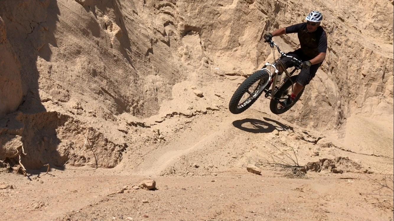 A mountain biker performing a jump over a sandy slope in a desert landscape. The rider is wearing a helmet and sports attire, capturing the essence of adventure and extreme sports. The backdrop features rocky terrain and a clear blue sky. Mariposa Fat Bike Trails mountain bike trail.