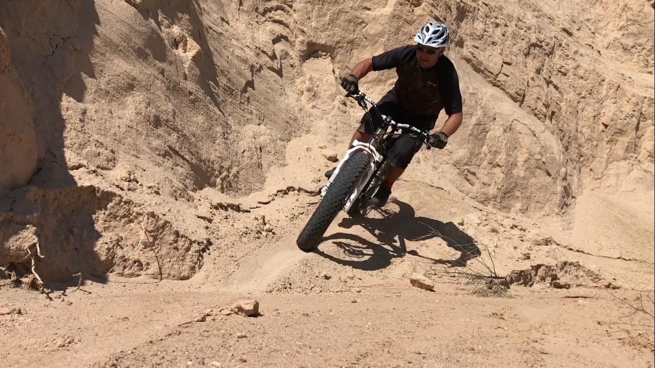 A mountain biker navigating a sandy slope on a trail, leaning into a turn. The cyclist is wearing a helmet, sunglasses, and casual biking attire, with a fat-tire bike designed for rough terrain. The background features steep, sandy hills. Mariposa Fat Bike Trails mountain bike trail.