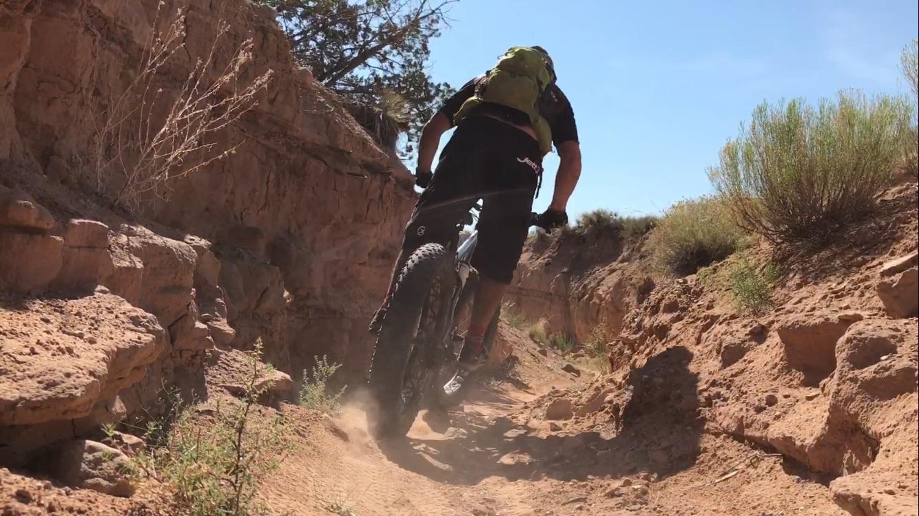 A mountain biker riding on a narrow dirt trail surrounded by rocky terrain and sparse vegetation, with a clear blue sky above. Dust rises from the bike's wheels as it navigates the path. Mariposa Fat Bike Trails mountain bike trail.