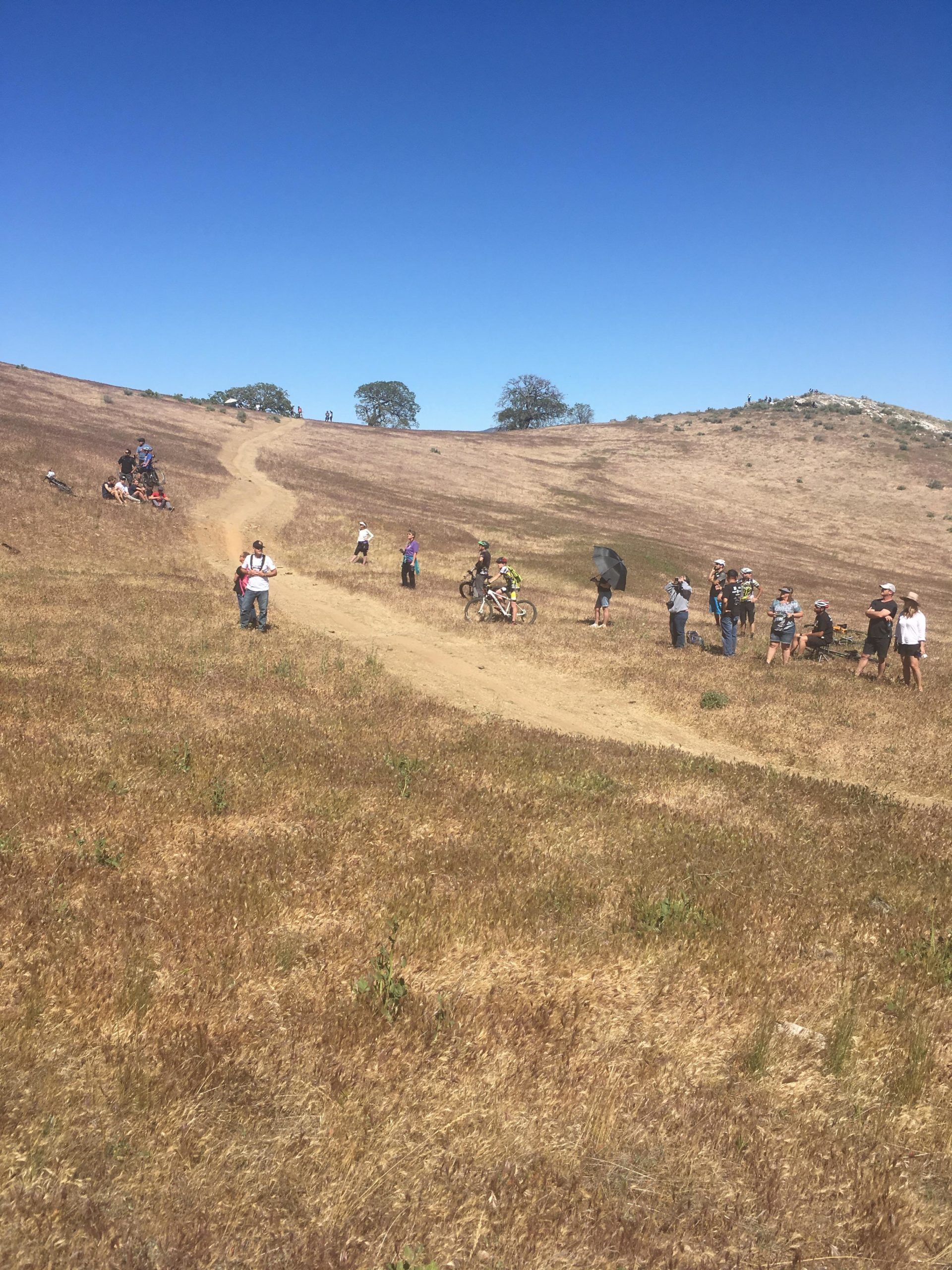 A group of people gathered on a dry, grassy hillside with a dirt path leading up the slope. Some individuals are sitting and resting, while others are standing and watching. A person on a bicycle is riding along the path, and a few trees can be seen in the background under a clear blue sky. NICA Loop mountain bike trail.
