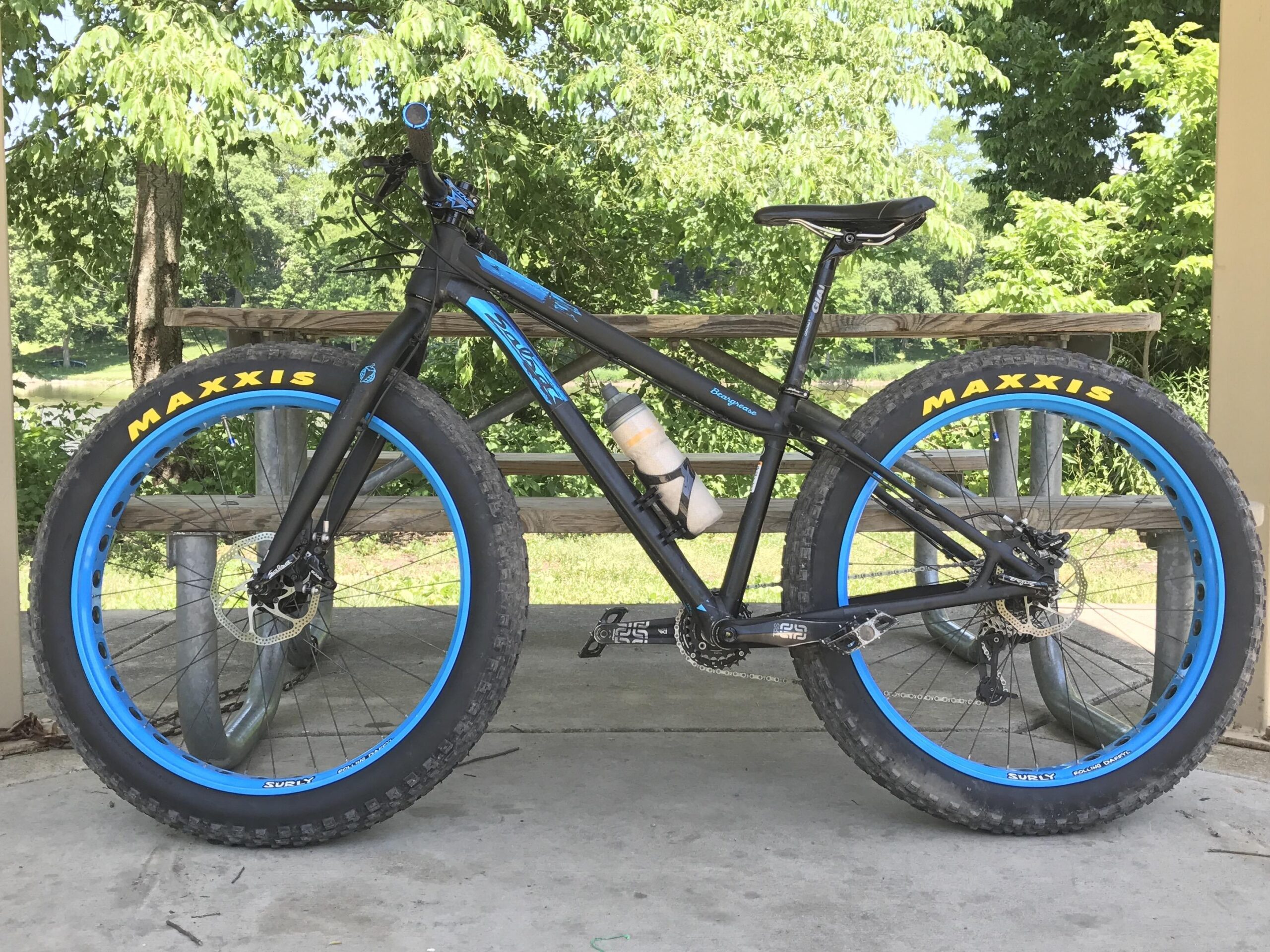 Salsa Beargrease: A black bicycle with blue accents and large tires is parked under a shelter surrounded by trees. The bike features wide, knobby tires labeled "MAXXIS" and has a water bottle holder attached. The background includes a glimpse of greenery, indicating a natural setting.