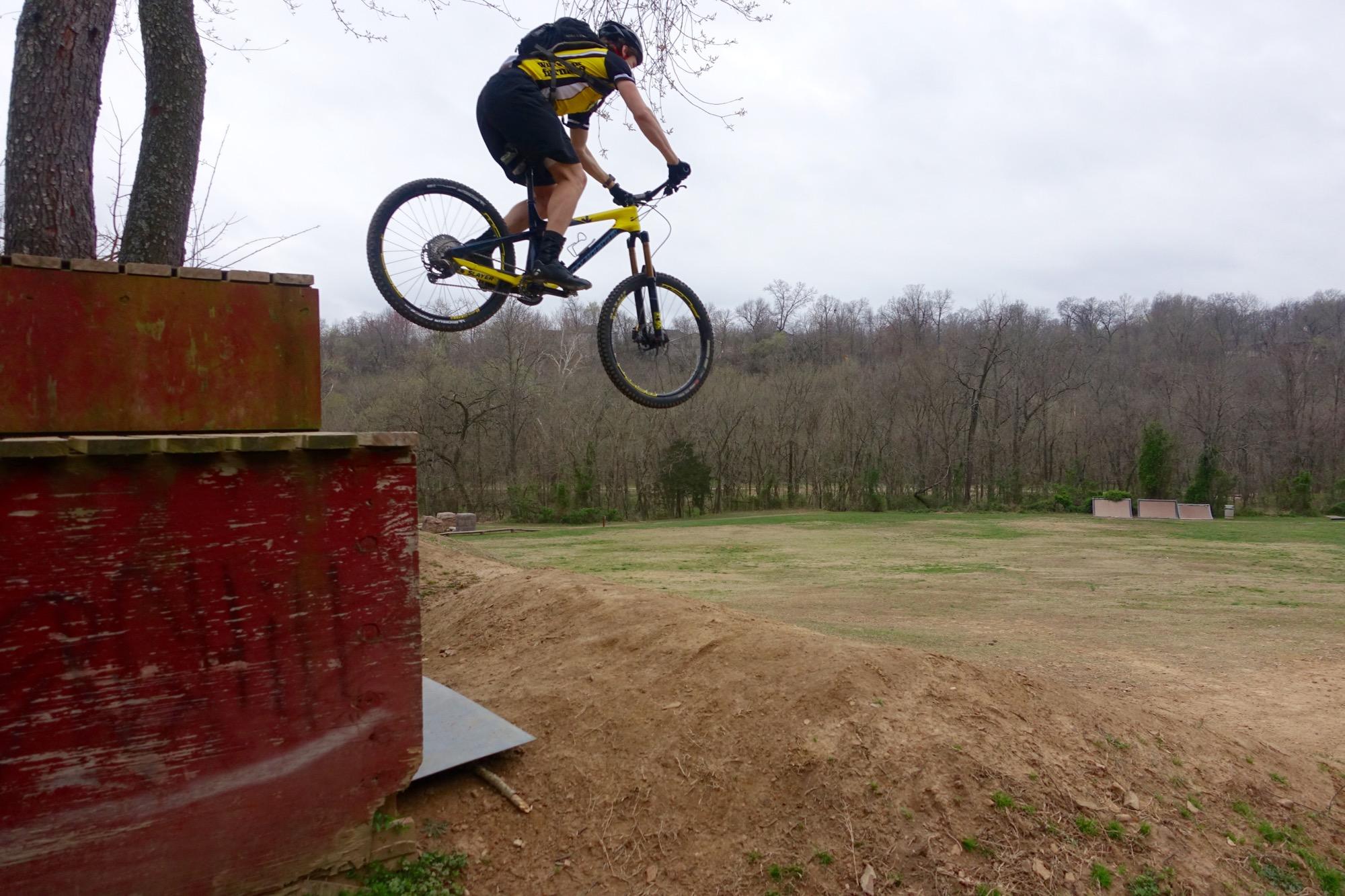 A cyclist wearing a yellow and black outfit is mid-air performing a jump off a ramp, with trees and a grassy landscape in the background. The scene captures the excitement of mountain biking in an outdoor setting. Slaughter Pen Trail mountain bike trail.