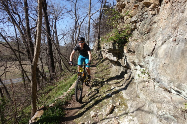 A mountain biker navigating a narrow, rocky trail surrounded by bare trees and a sunny blue sky. The rider is wearing a helmet and sunglasses, dressed in a black shirt and turquoise shorts, as they pedal along the rugged path.