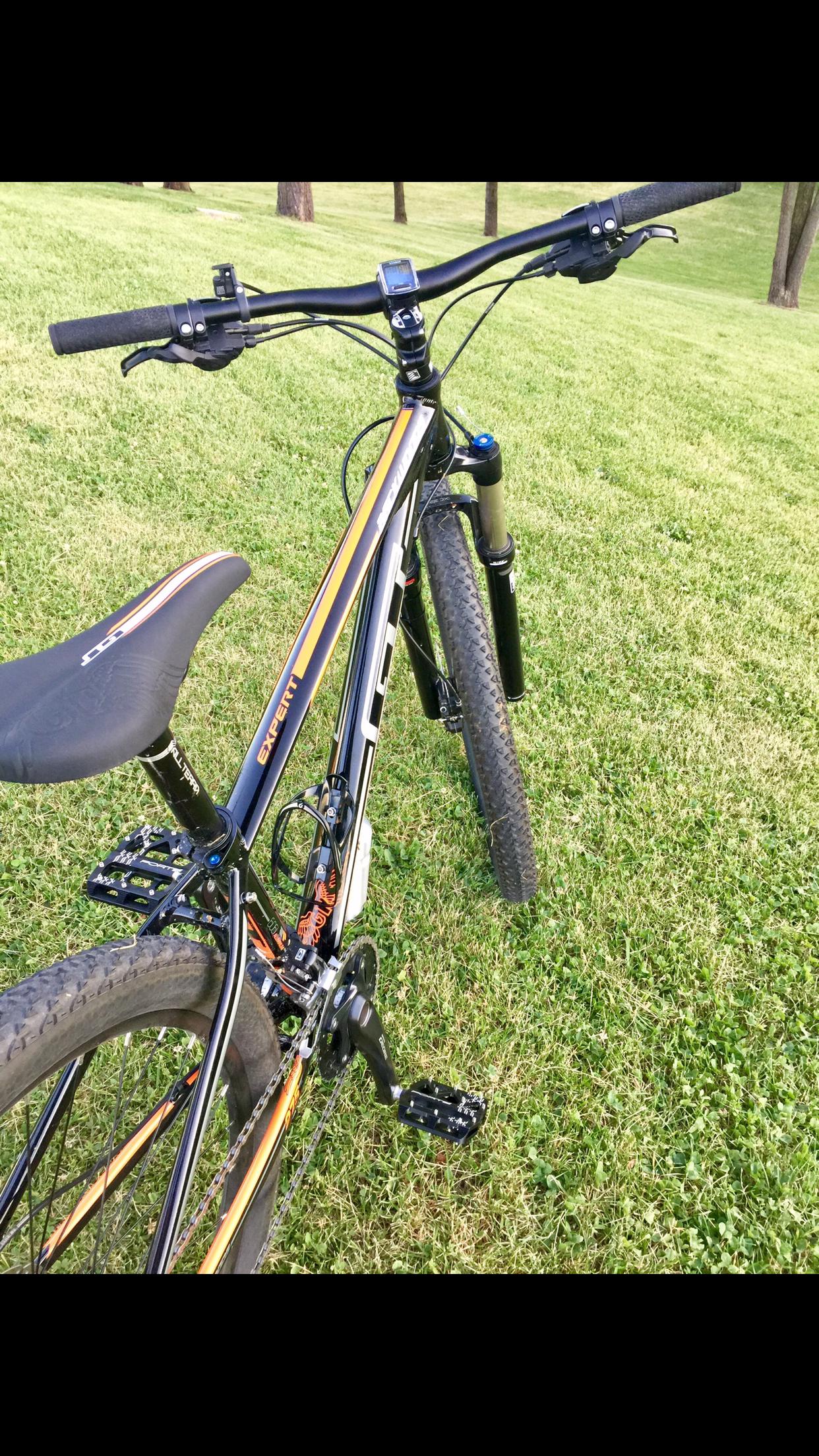 GT Backwoods: A close-up view of a mountain bike placed on green grass, showcasing the handlebars, wheels, and pedals. The bike features a black and orange frame with visible gears and a saddle designed for comfort. In the background, there are trees and a slight incline in the terrain.